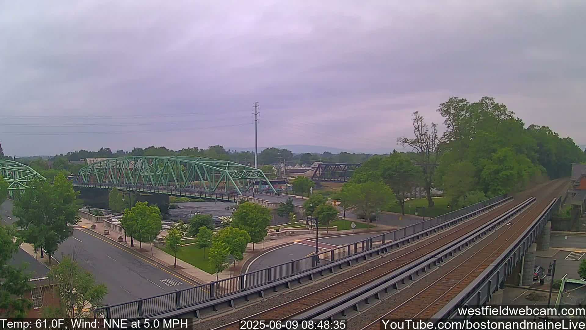 A view of a town on an overcast day, featuring two bridges over a river, train tracks elevated on a bridge, and lush green trees surrounding the area.