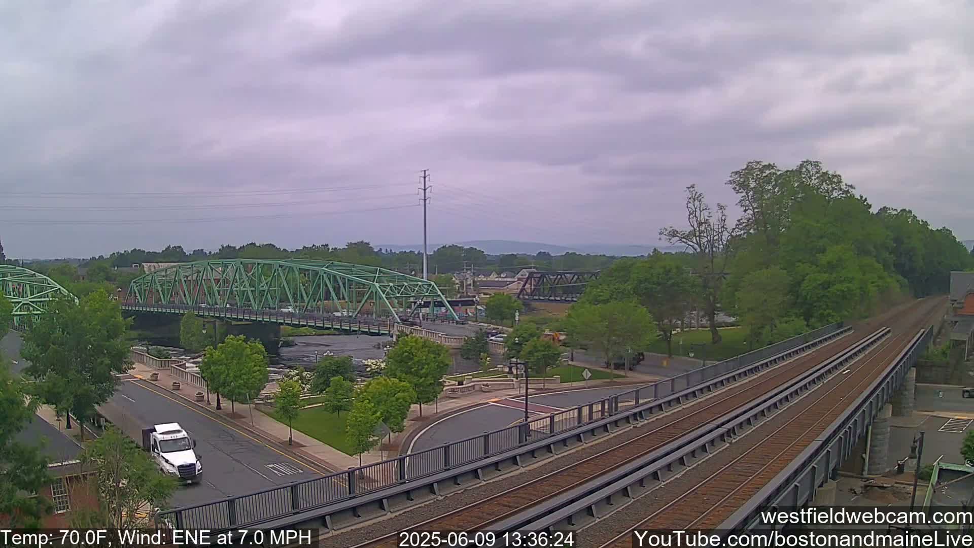 A partly cloudy day reveals a scene of two bridges spanning a river, with train tracks and a road visible below, surrounded by green trees and landscaping.