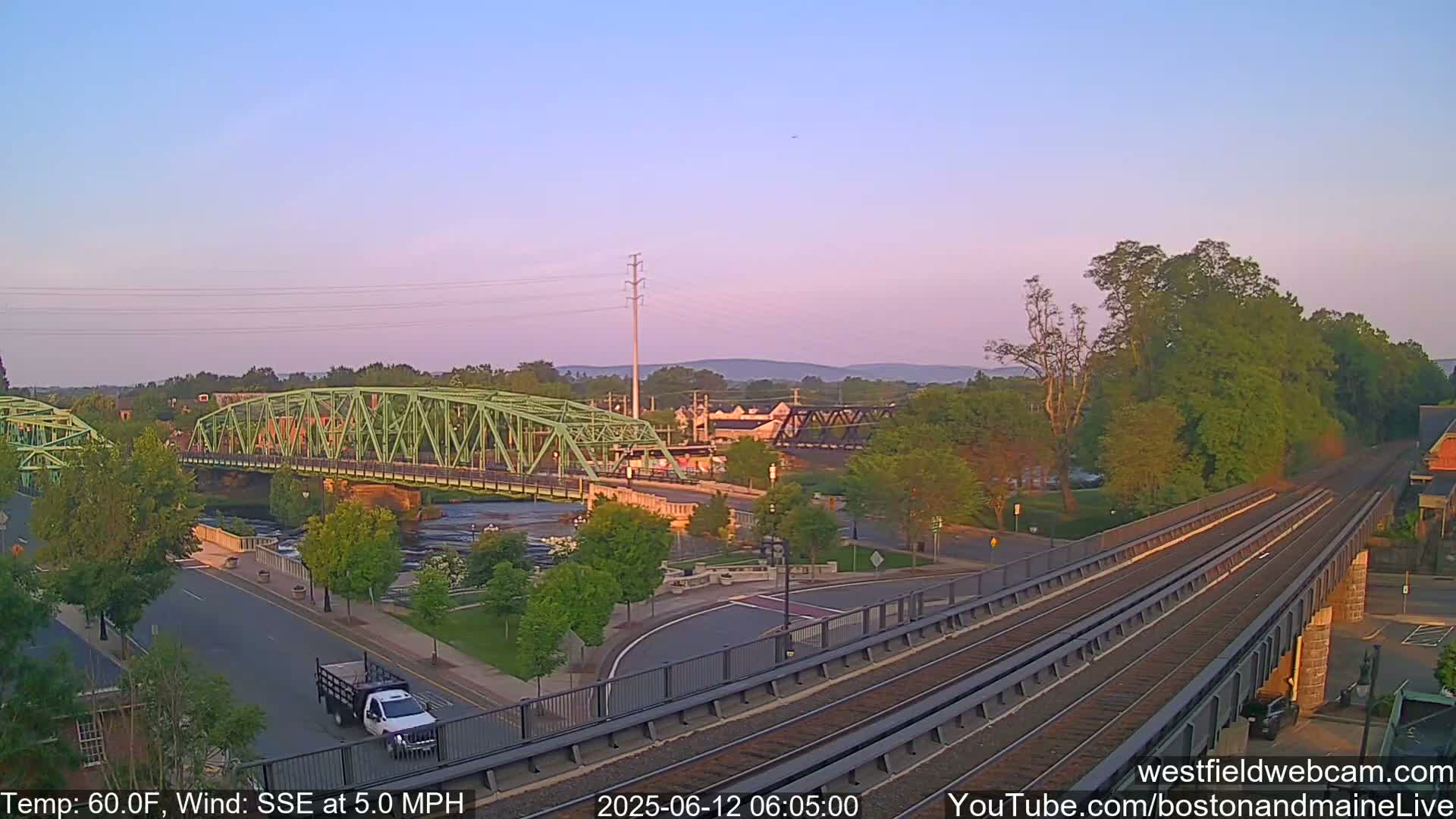 A pale pink sunrise illuminates a town scene featuring two bridges over a river, train tracks, and lush green trees.