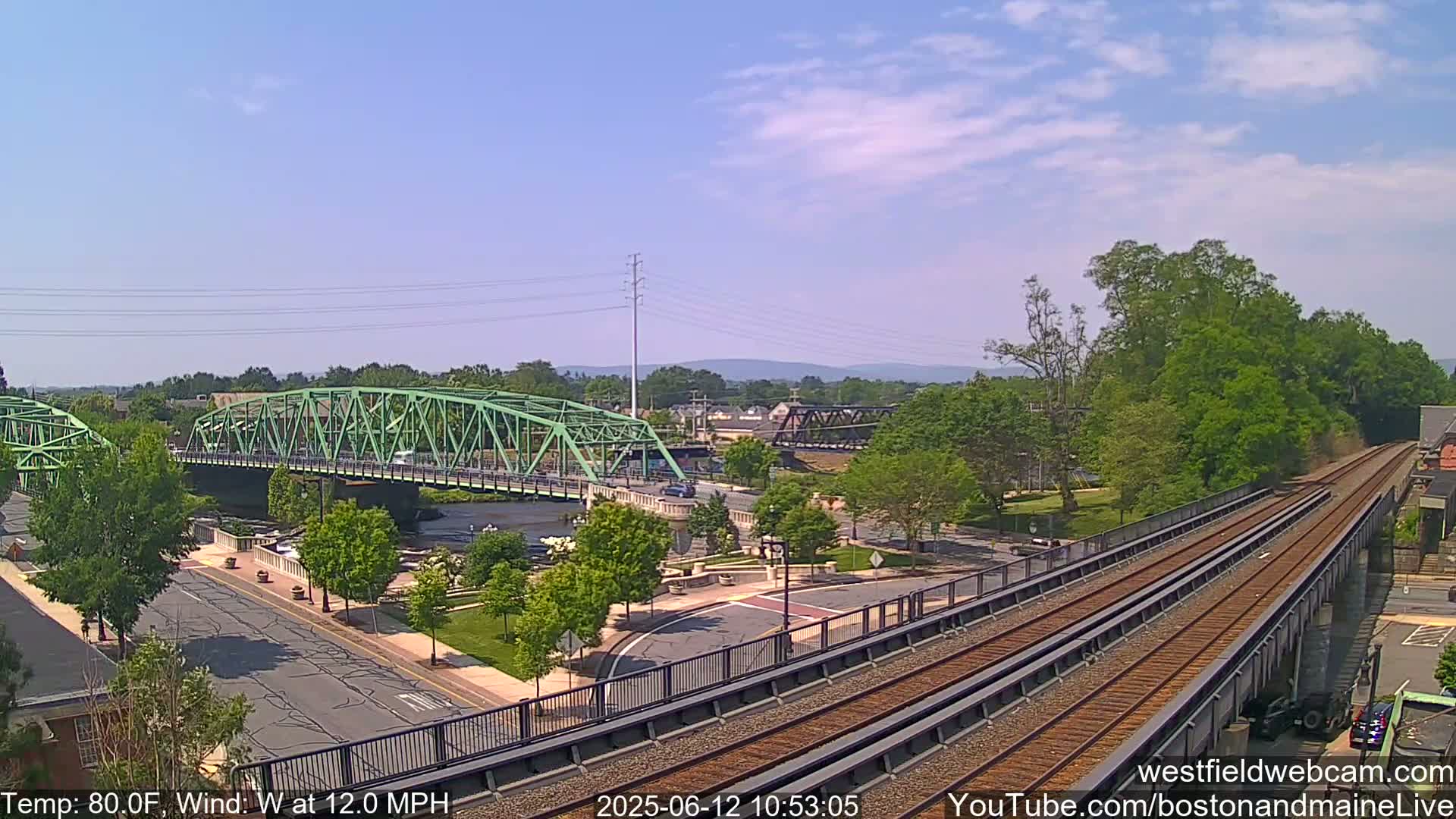 A view of a town on a sunny day with a green bridge over a river and train tracks in the foreground, with a light wind at 12 mph and a temperature of 80°F.
