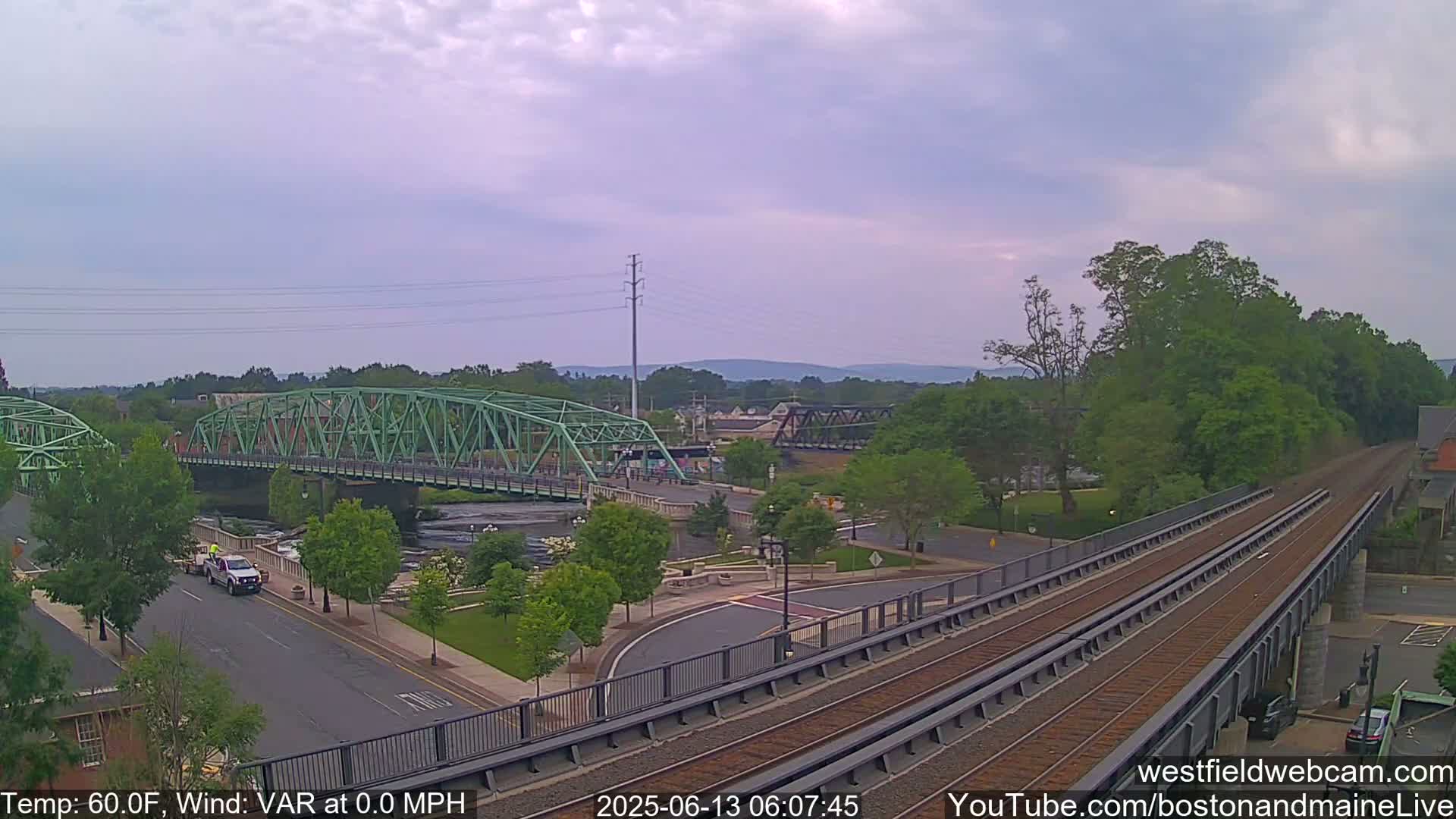 A light-green bridge spans a river, with train tracks and a road intersecting nearby under a partly cloudy sky.