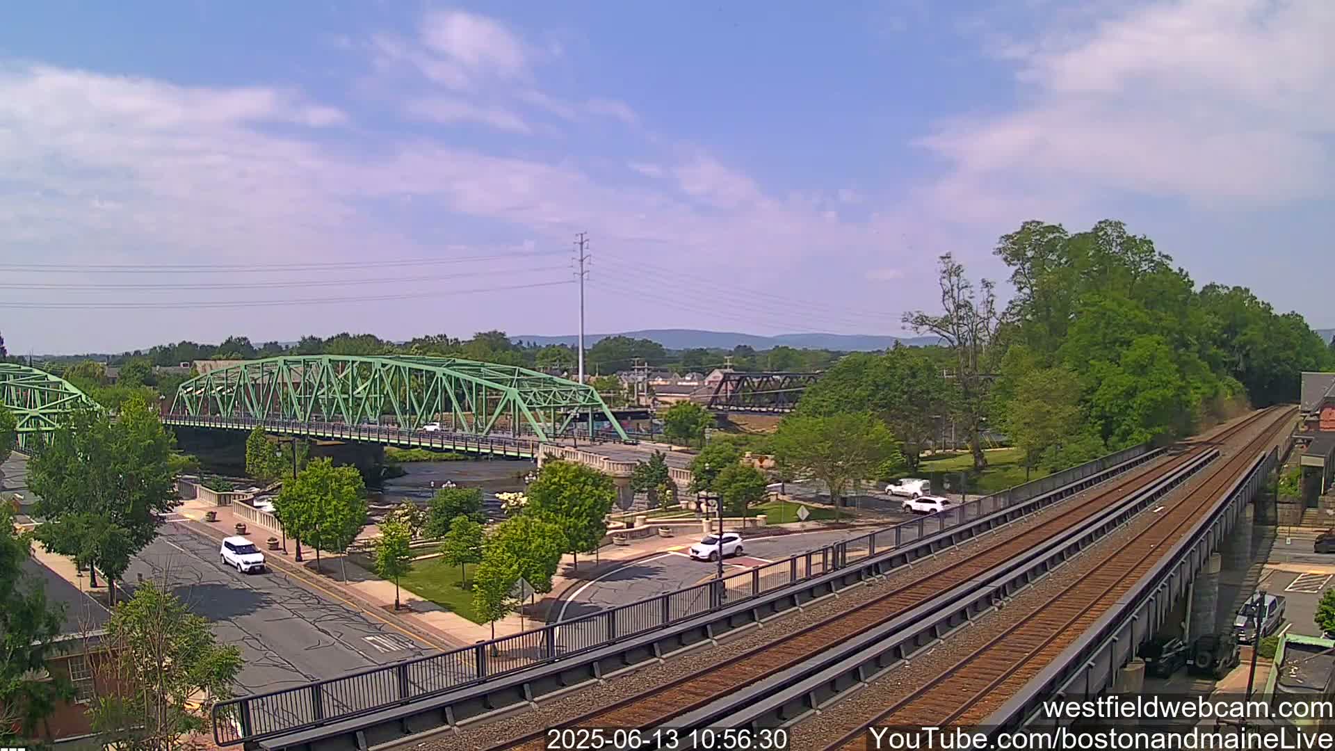 A high-angle view on a sunny day shows a green bridge over a river, a train track elevated above a roadway, and several cars at an intersection, all surrounded by lush green trees.