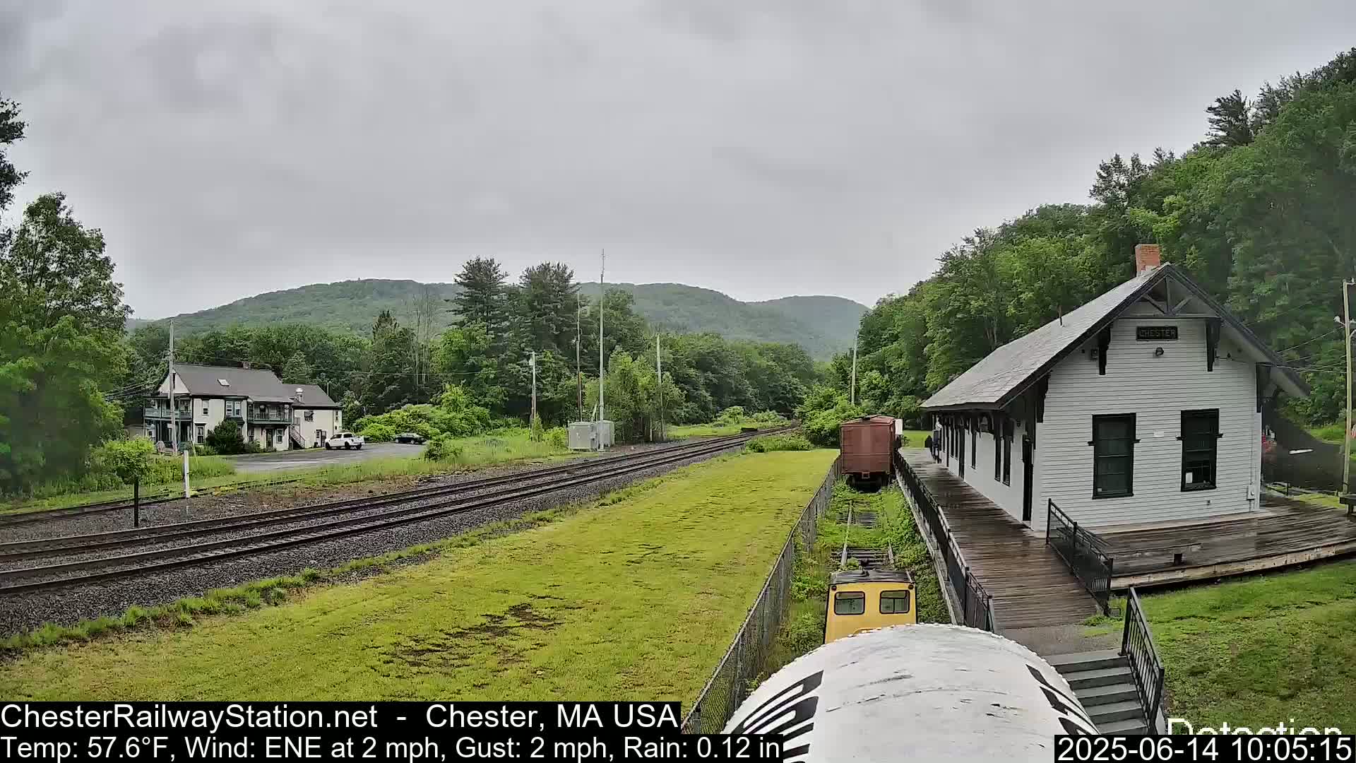 A train approaches a small, white train station on a rainy day, with green hills and trees in the background.