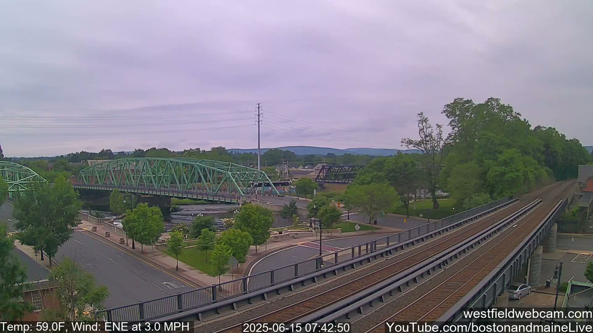 A green bridge spans a river in a town, with train tracks elevated alongside it under an overcast sky, the temperature at 59°F and a northeasterly wind at 3.0 mph.