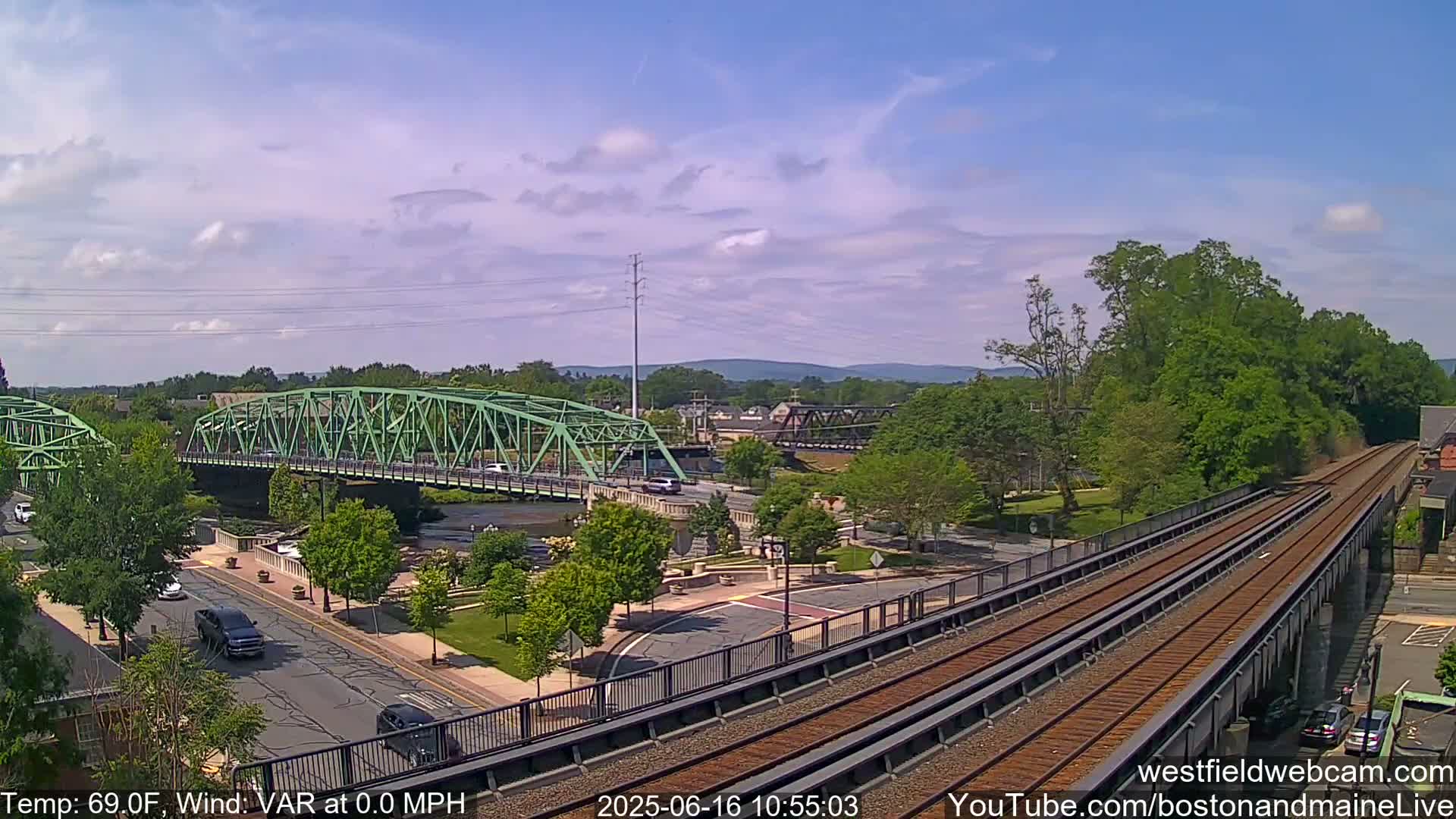 A green bridge spans a river under a partly cloudy sky, with train tracks and roadways visible below, all set against a backdrop of lush green trees and distant hills.