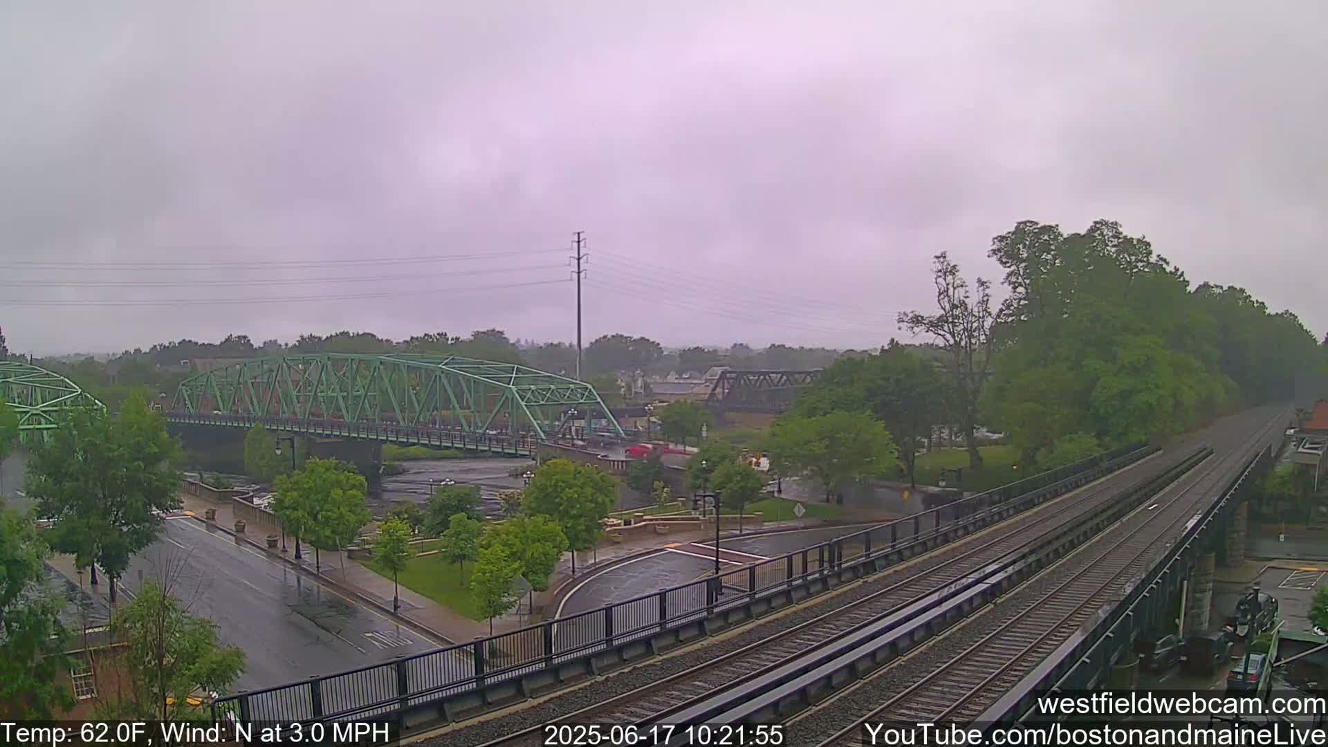 A light rain falls on a town scene featuring two bridges over a river, a roadway, and train tracks, with lush green trees lining the riverbanks.