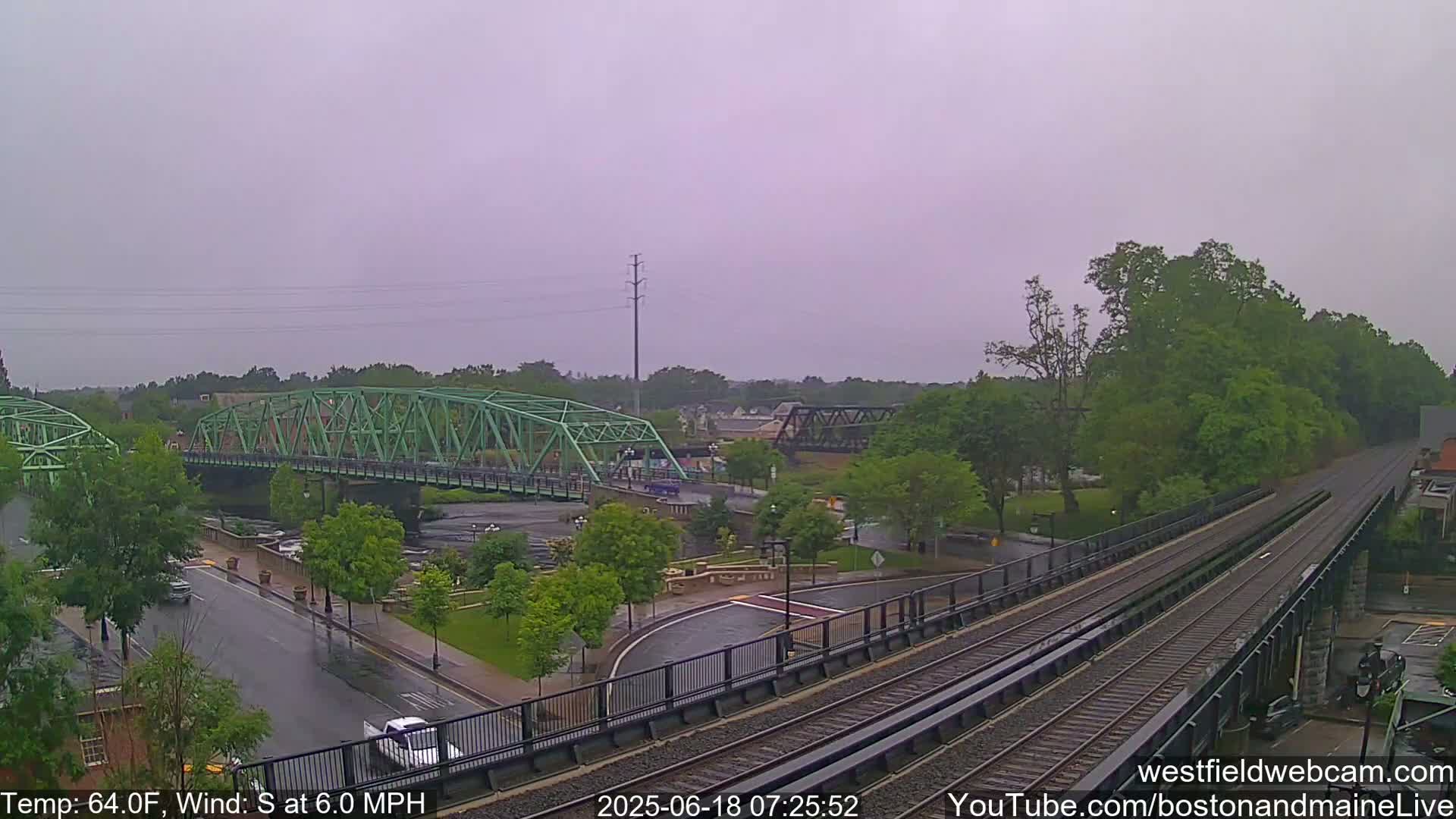 A light-green bridge spans a river, with train tracks and a wet roadway visible underneath a cloudy sky.