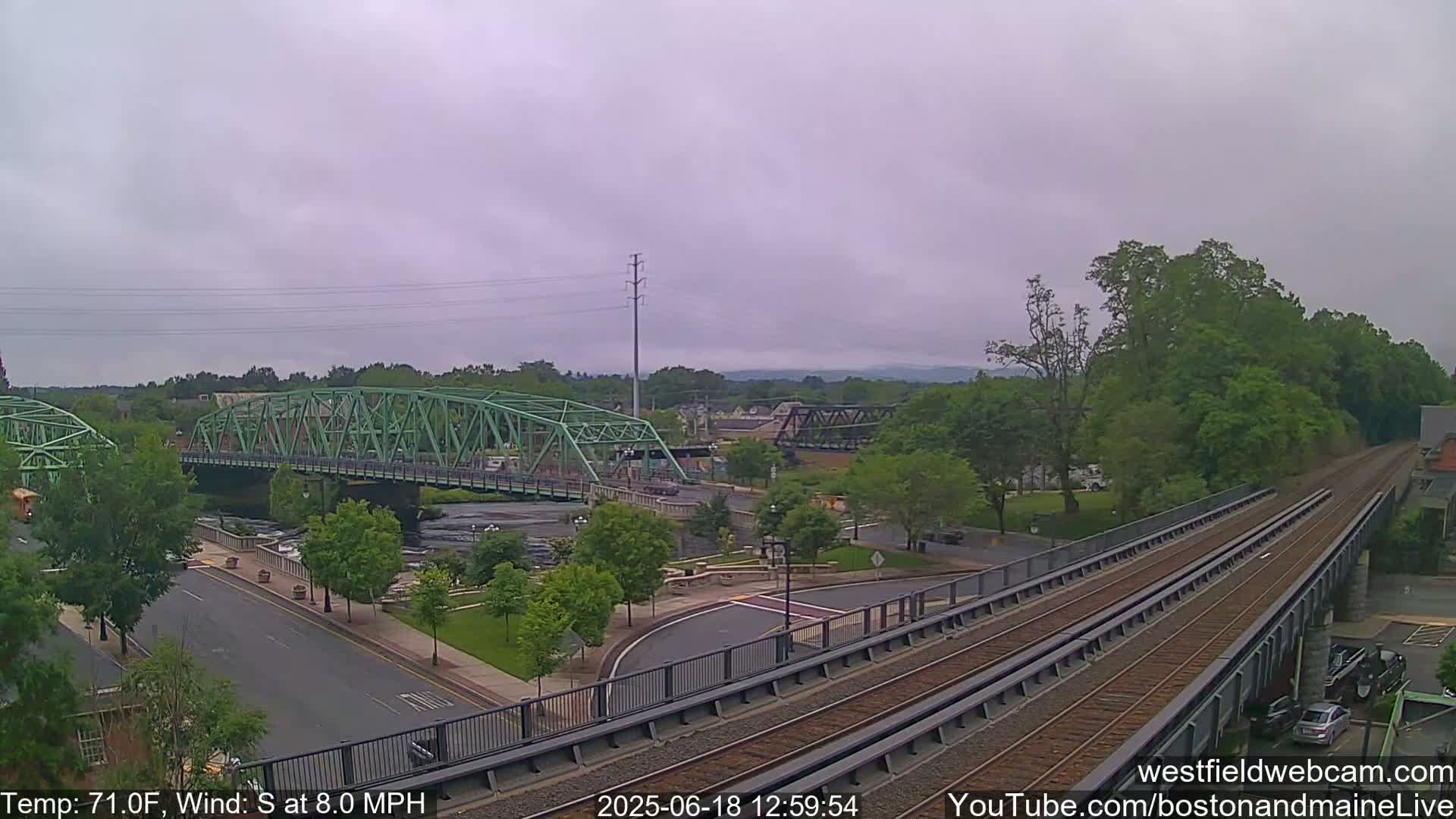 A view of a town with two bridges over a river, one green and one dark metal, under an overcast sky with a south wind.