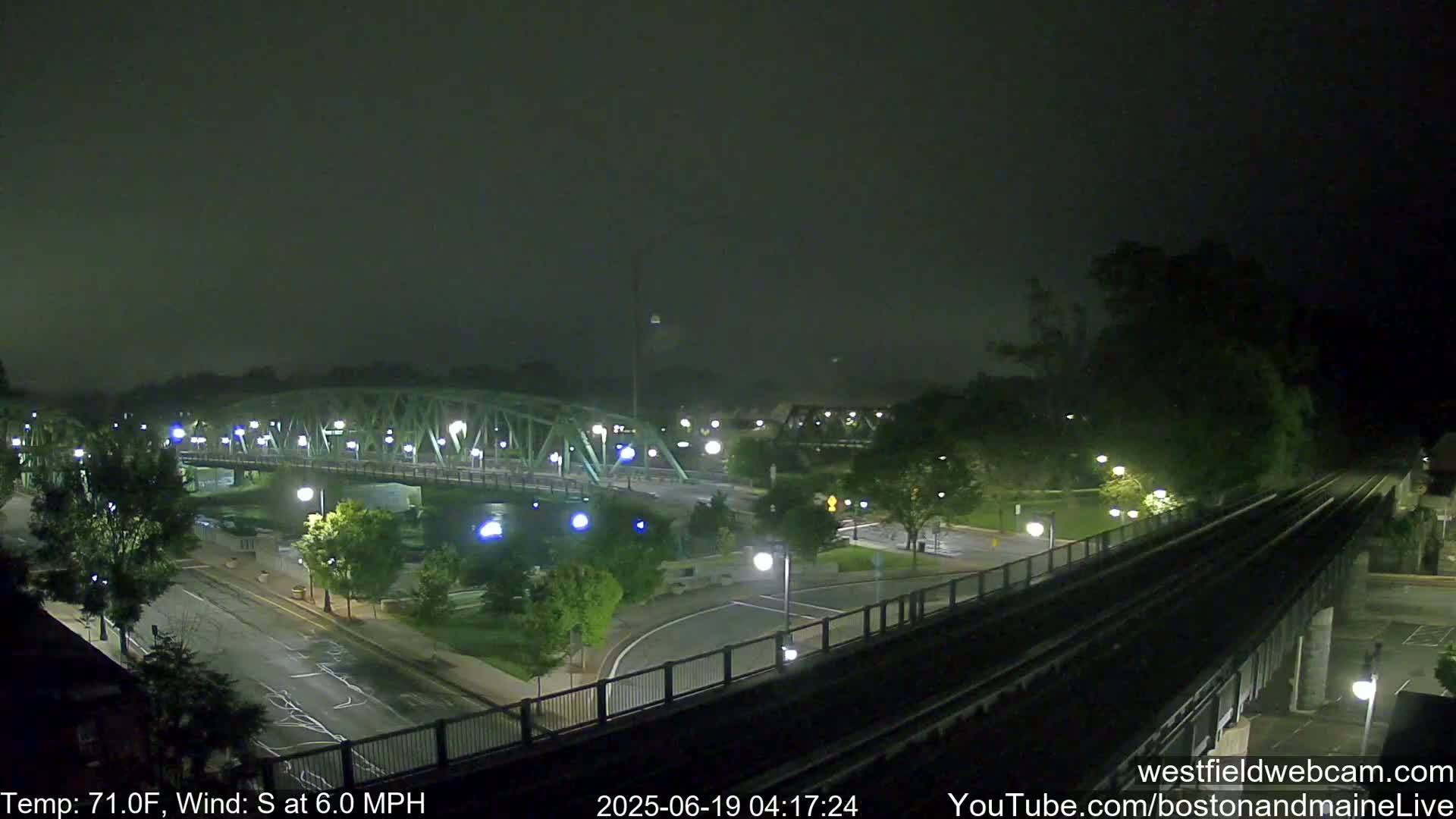 A nighttime view of a bridge and road intersection, with a railway track elevated alongside, under hazy conditions and a south wind at 6 mph.