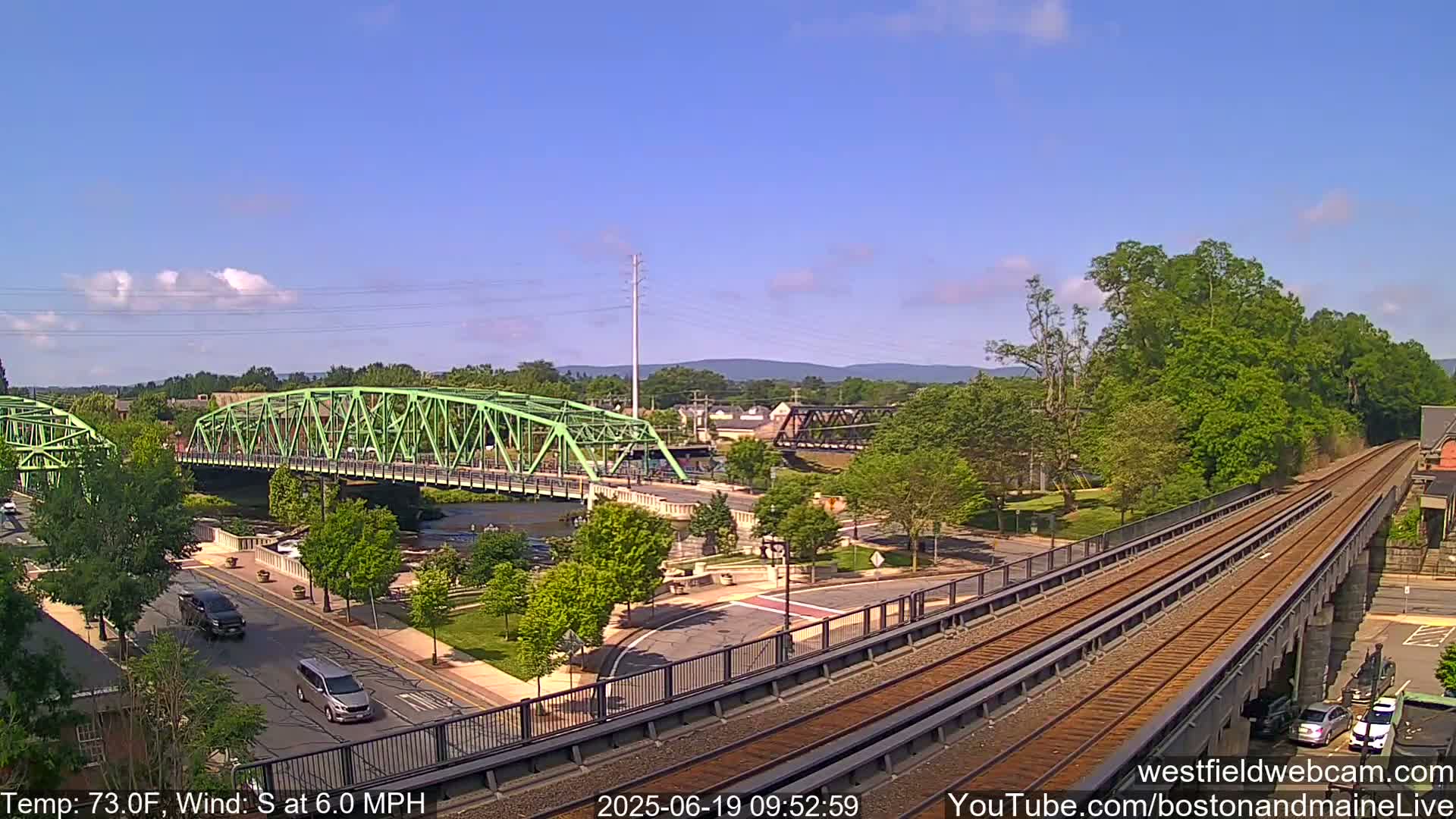 A green bridge spans a river, with a train track elevated alongside a road, all under a mostly clear sky and a temperature of 73 degrees Fahrenheit, with a south wind at 6 mph.