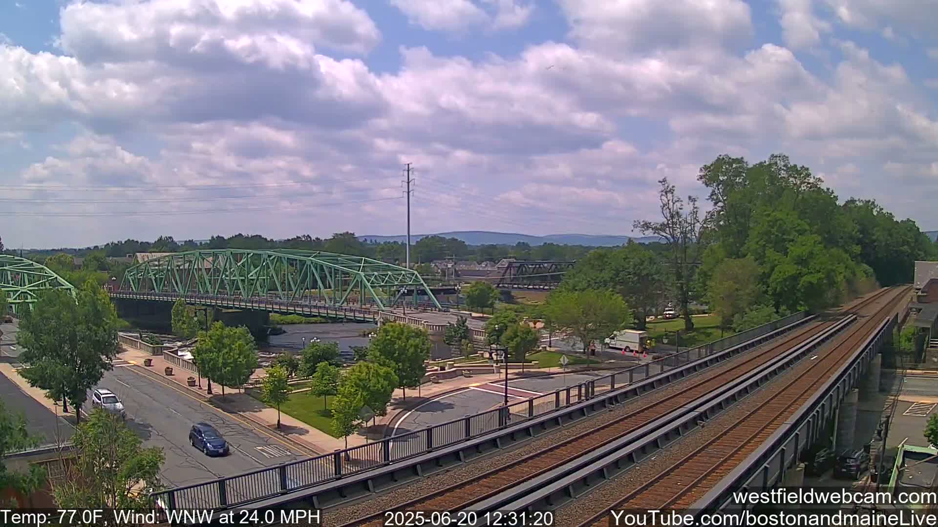 A partly cloudy day with a WNW wind at 24 mph shows a view of a town with a green bridge over a river, a train track, and cars on a road below.