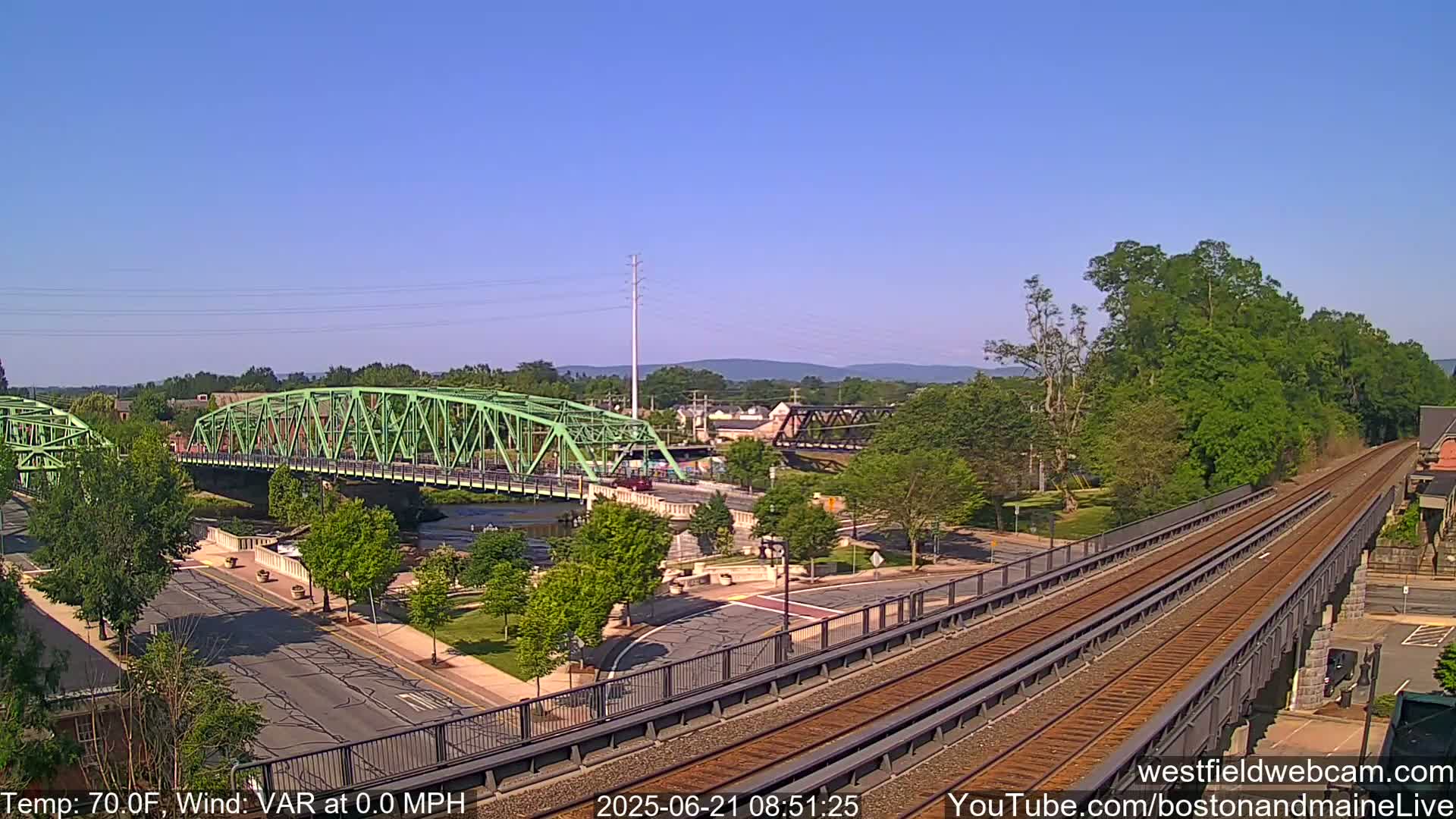 A view of a town on a clear, sunny day, showing a river with two bridges, one green and one dark metal, and train tracks crossing over a road.