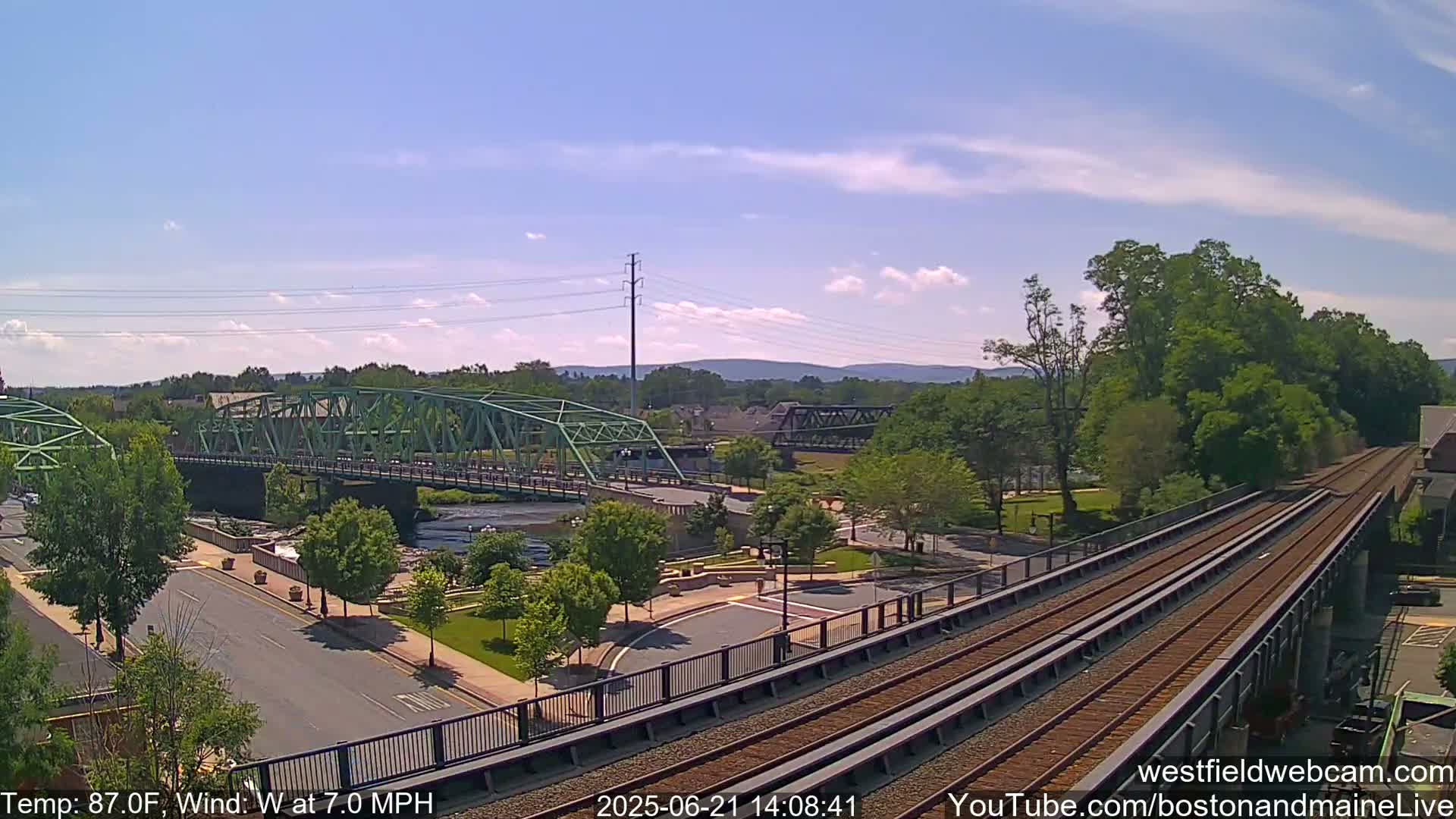 A high-angle view shows a town on a sunny day with a river, two bridges, and train tracks under a mostly clear sky.