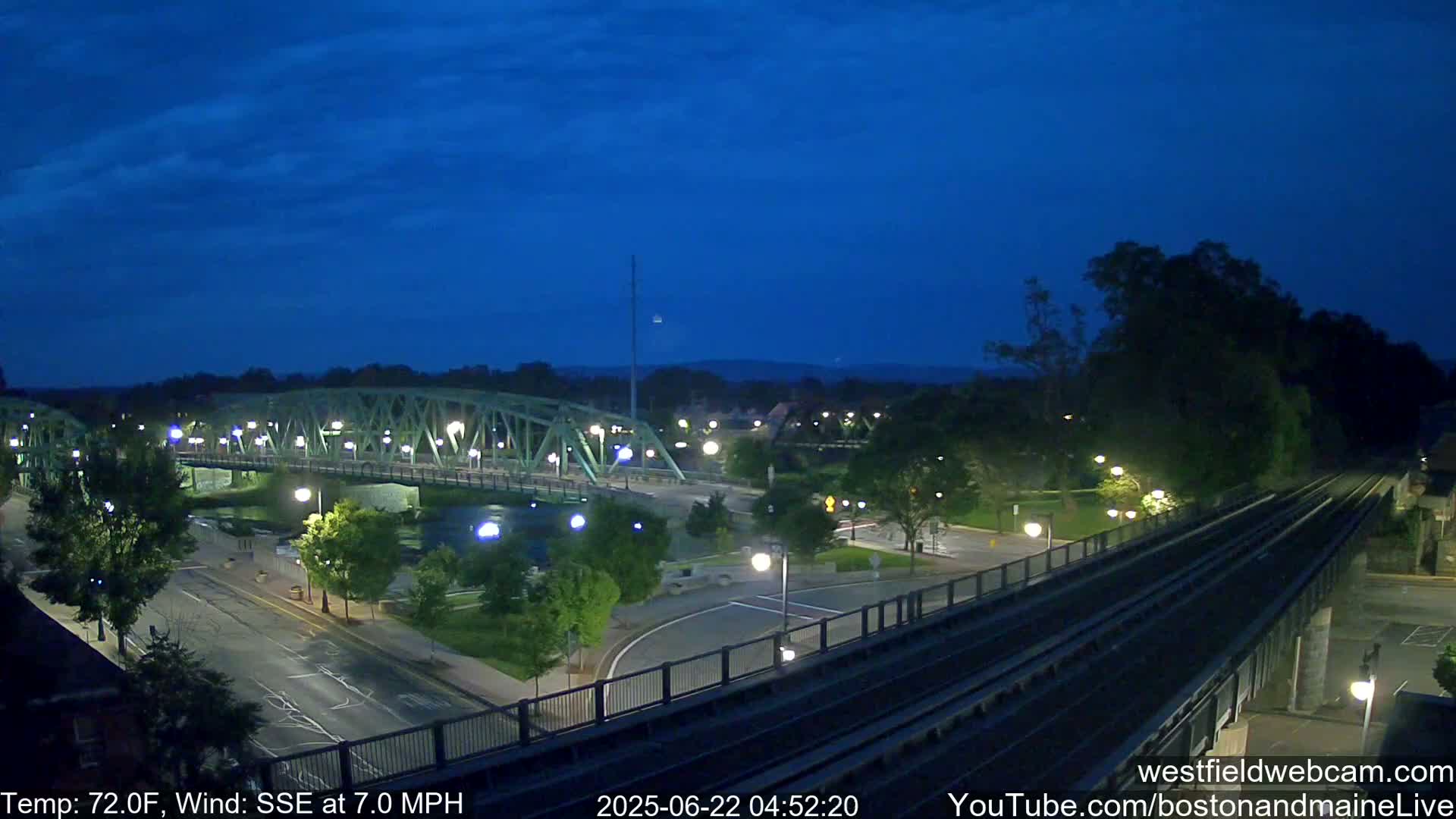 A nighttime view shows a bridge over a river, a road, and a train track, under a dark blue, clear sky.