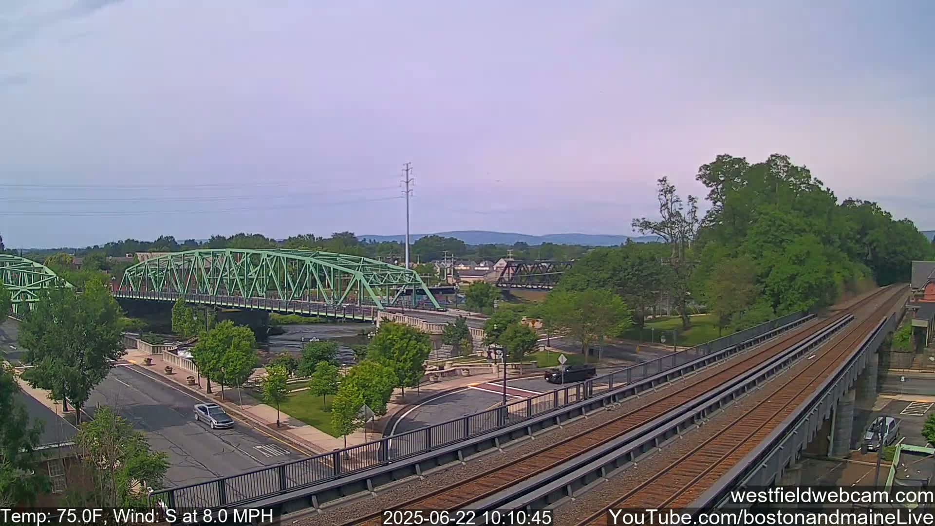 A view from an elevated train track shows a green bridge over a river, a roadway, and lush green trees under a partly cloudy sky, with a temperature of 75 degrees Fahrenheit and a south wind at 8 miles per hour.