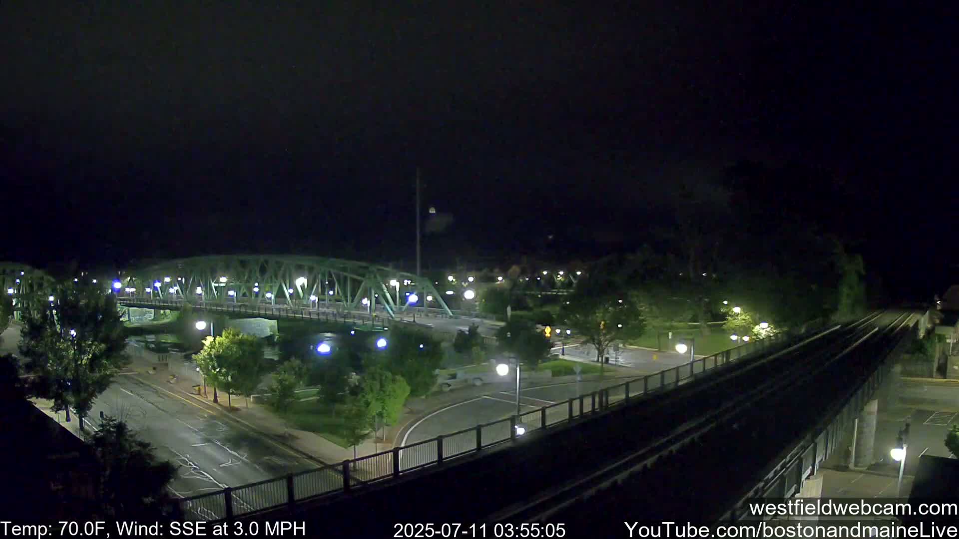 A nighttime view of a bridge over a river, with a road and train tracks intersecting below, under a clear, dark sky with a light wind.
