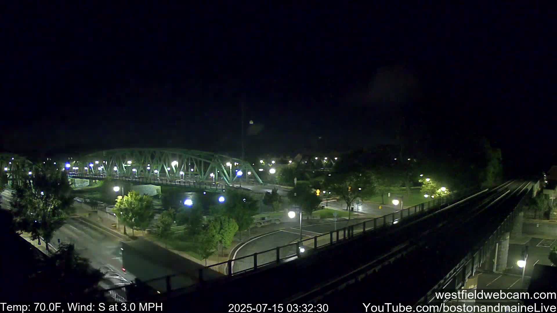 A nighttime view of a green metal bridge over a waterway, with a road and park below, under a dark, clear sky with a slight south wind.