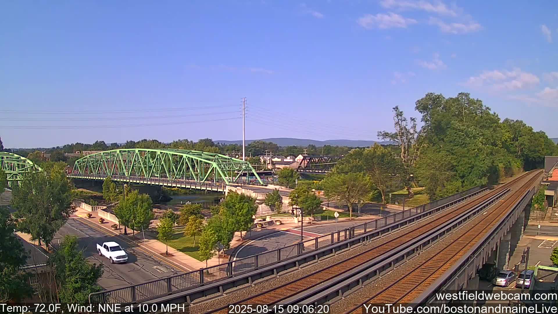A rainy view of a river, two bridges, a roadway, and train tracks, with green trees and overcast skies.