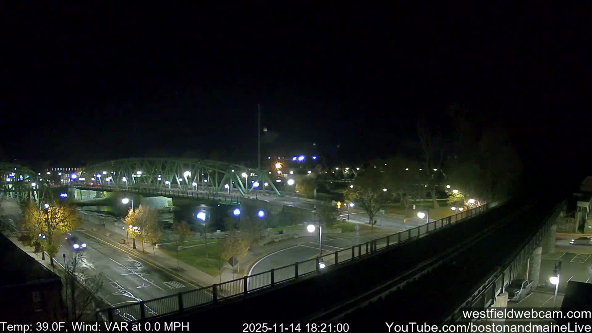 A clear and calm 39-degree Fahrenheit night presents a cityscape centered around a large green truss bridge crossing a river, with illuminated roads below showing signs of vehicle traffic and numerous bright streetlights.