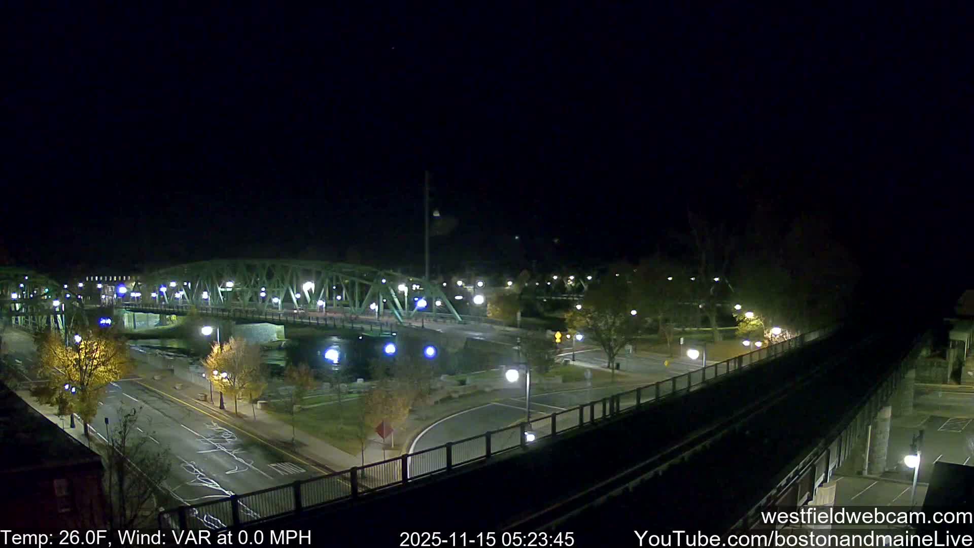 A well-lit city scene at night features a green truss bridge over a dark river, a wet road, and elevated train tracks, all illuminated by numerous streetlights under clear, cold conditions.