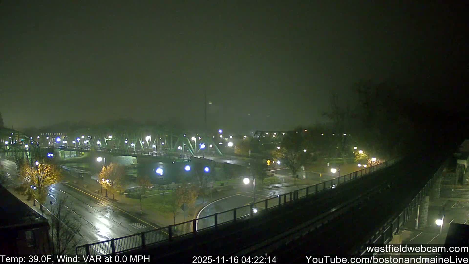 An elevated night scene shows city roads and green truss bridges illuminated by streetlights, with their reflections shimmering on wet pavement under a hazy sky.