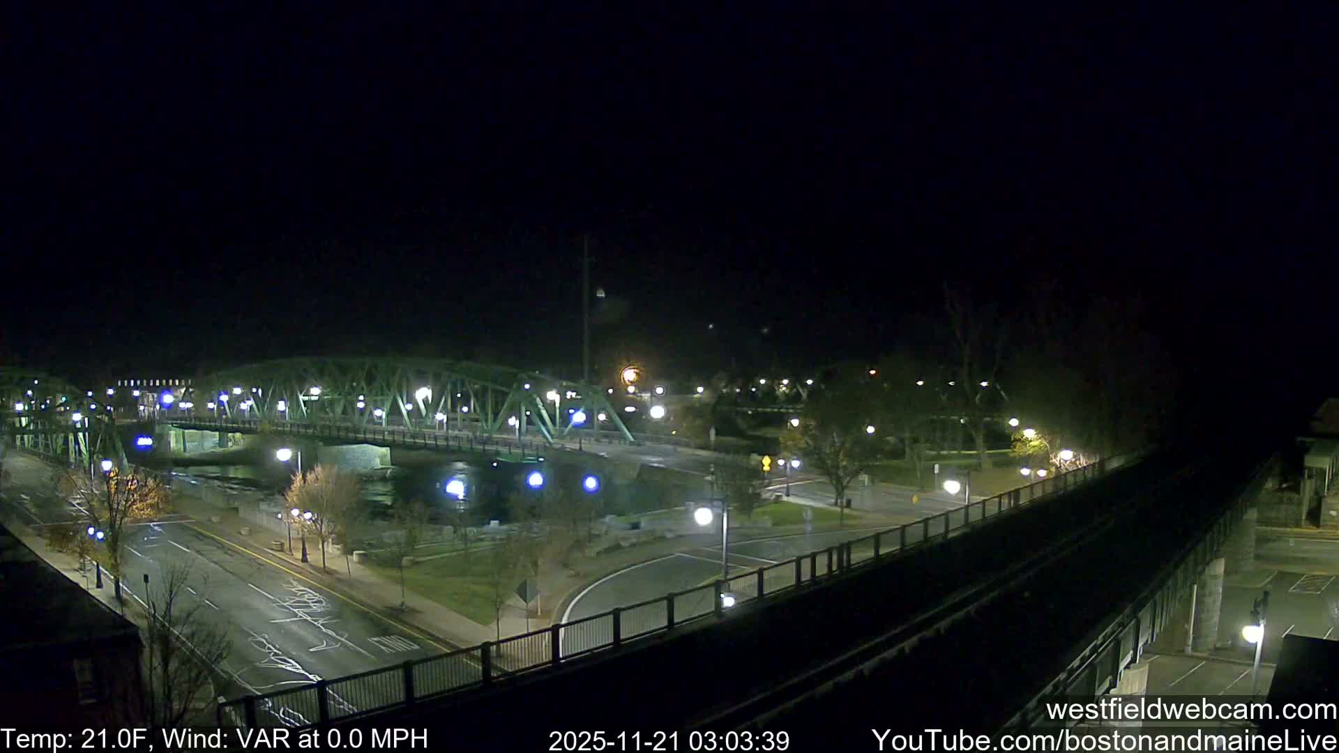 A clear night view captures a city scene with a prominent green truss bridge, brightly illuminated by numerous streetlights, spanning a river alongside roads, bare trees, and an elevated track in the foreground.