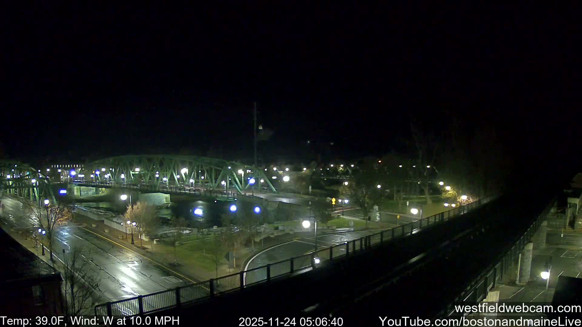 A nighttime aerial view captures a vibrant green truss bridge spanning a river, with wet roads and pathways reflecting numerous streetlights under a clear dark sky, suggesting recent rain.
