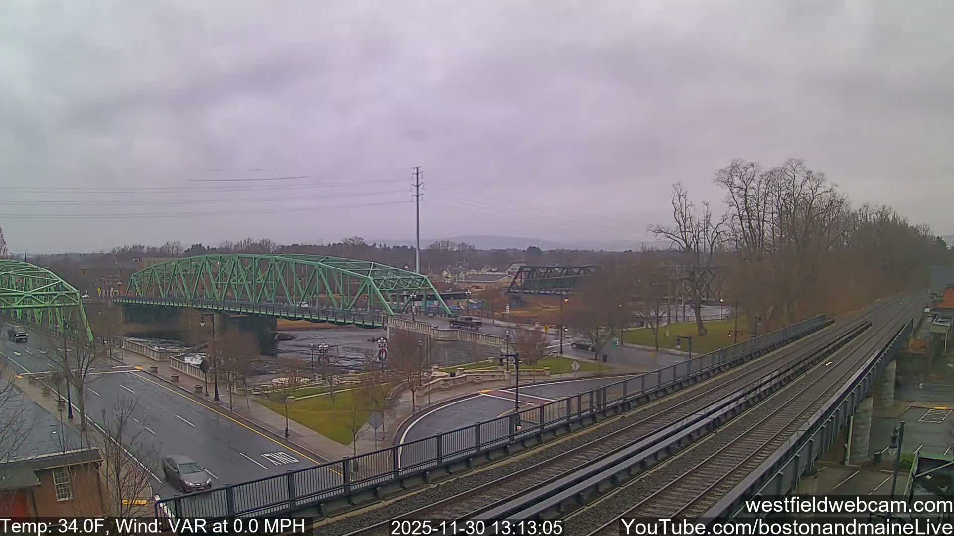 A clear night reveals a large green truss bridge spanning a river, flanked by illuminated city streets, some bare trees, and an elevated railway structure on the right.