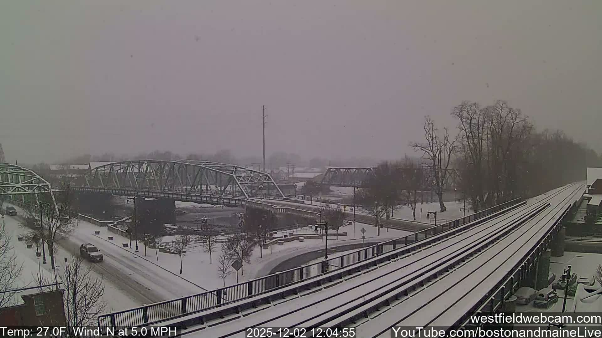 An elevated view reveals a winter landscape blanketed in snow under an overcast sky, showcasing multiple truss bridges over a river and streets, with bare trees, moving vehicles, and snow-covered railroad tracks extending into the distance.