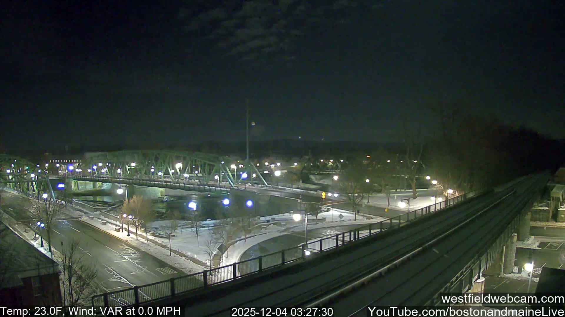 The image displays a snowy urban landscape at night, featuring a large green truss bridge crossing a river, illuminated by numerous streetlights, with roads, bare trees, and an elevated railway line visible under a dark, partly cloudy sky.
