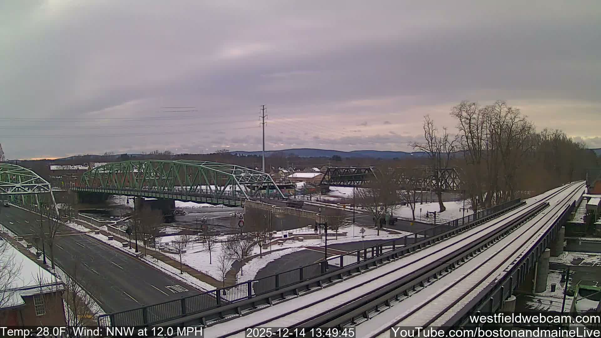 The image displays a snowy urban landscape at night, featuring a large green truss bridge crossing a river, illuminated by numerous streetlights, with roads, bare trees, and an elevated railway line visible under a dark, partly cloudy sky.