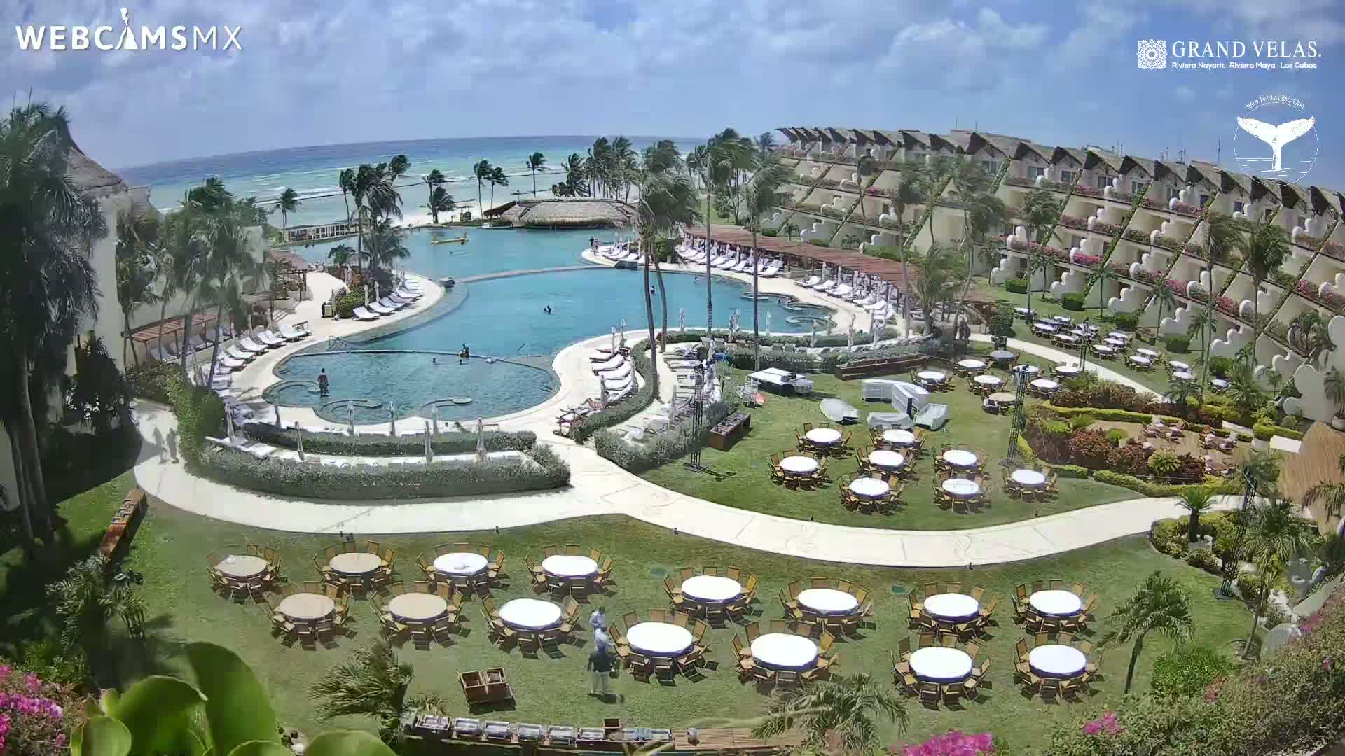 An aerial view of a resort with a large pool, multiple buildings, palm trees, and numerous tables set up on a grassy area, all under a sunny sky with a clear ocean in the background.