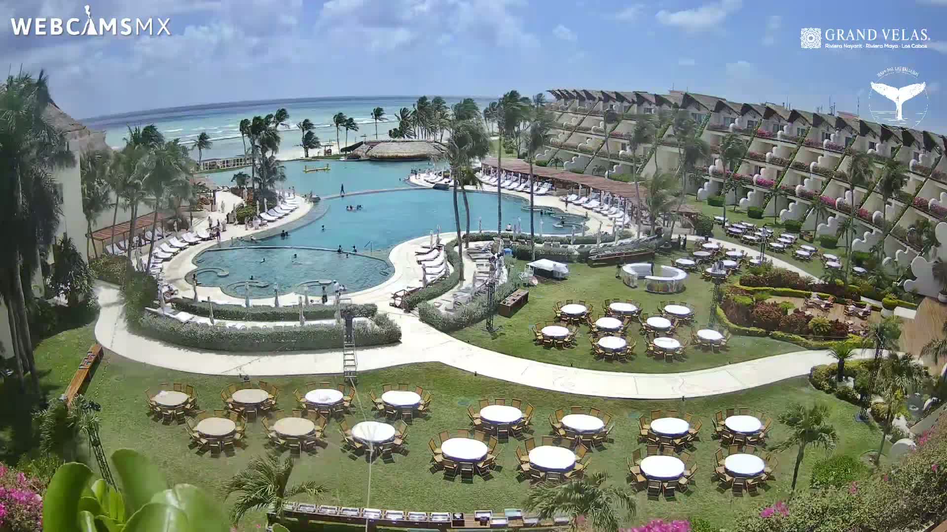 An aerial view of a resort on a sunny day shows a large pool, multiple buildings, and numerous tables set up on a grassy area near the beach.