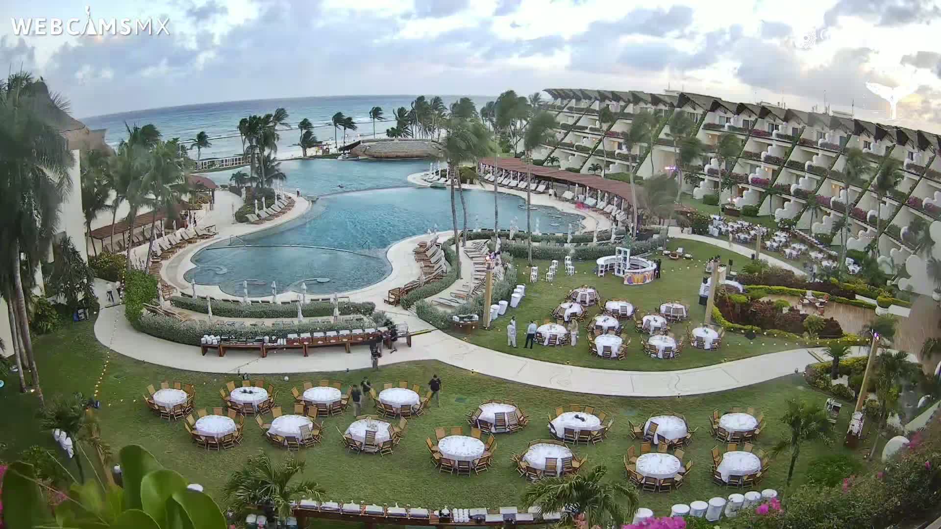 An outdoor resort scene shows a large pool, a multi-story building, and numerous tables set for an event on a grassy area, all under a partly cloudy sky with a windy ocean in the background.