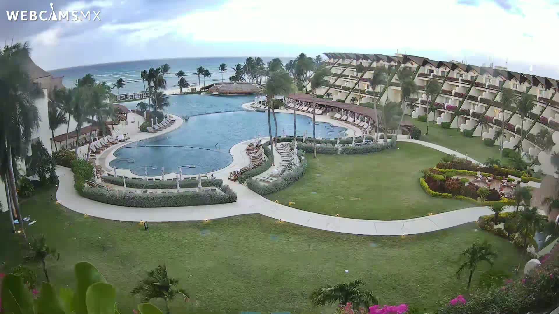 An aerial view shows a resort with a large, winding pool surrounded by buildings and lush green landscaping, overlooking a partly cloudy ocean.