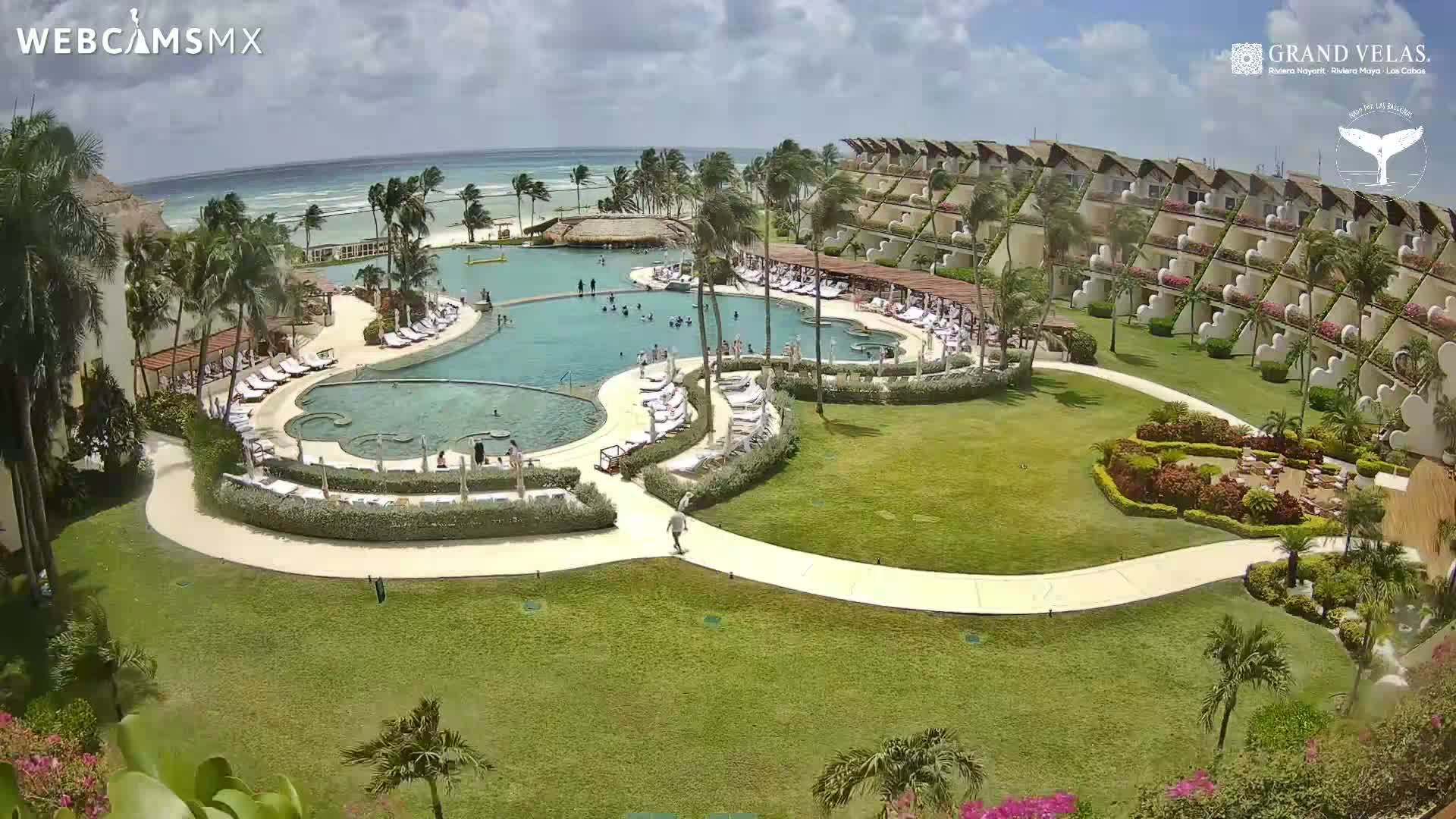 An aerial view shows a resort pool area with many people swimming and lounging, surrounded by lush landscaping, buildings, and a beach with a partly cloudy sky.