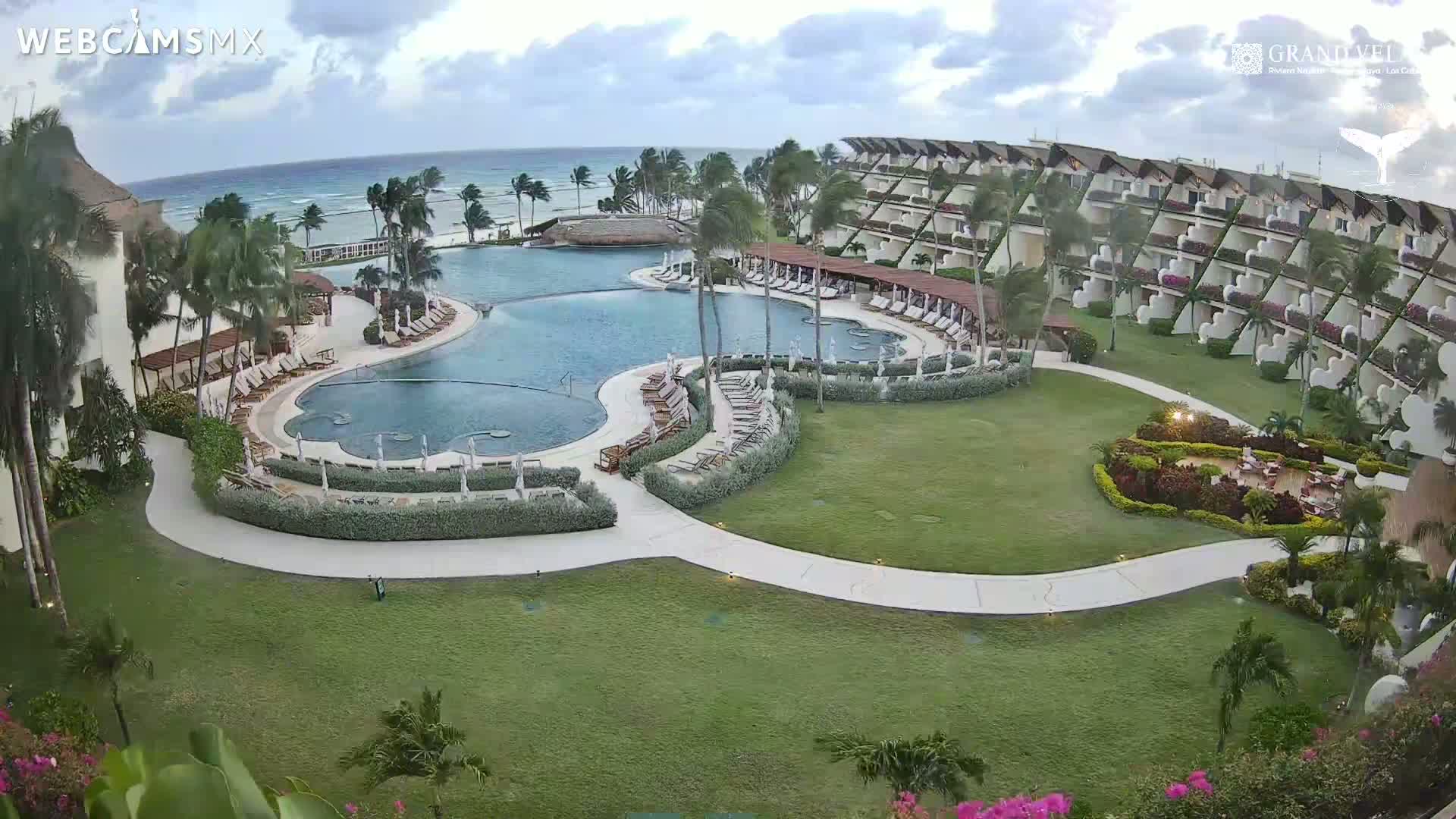An aerial view of a resort with a large, curved pool, surrounding landscaping, and a multi-story building overlooking a calm ocean under a partly cloudy sky.