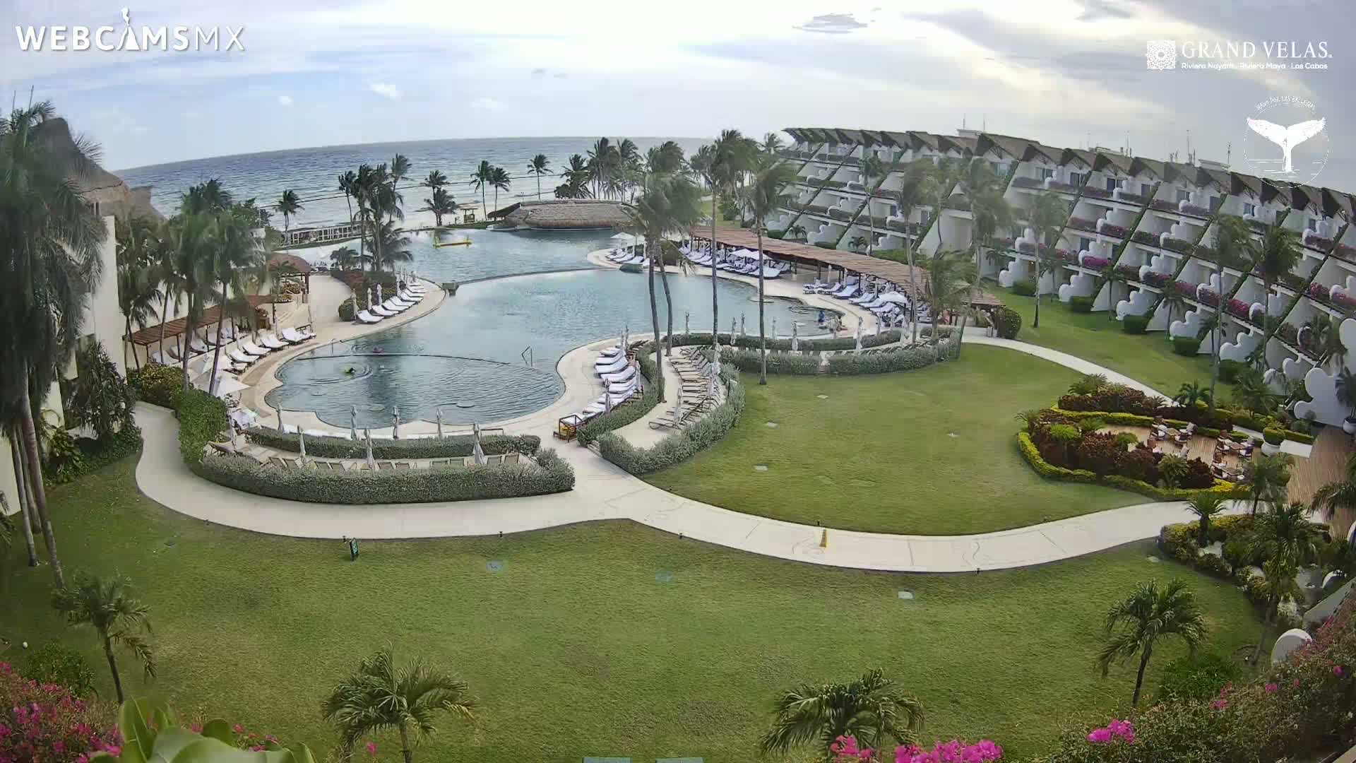 An aerial view shows a resort with a large, curved pool surrounded by lounge chairs and lush landscaping, situated next to a calm ocean under a partly cloudy sky.