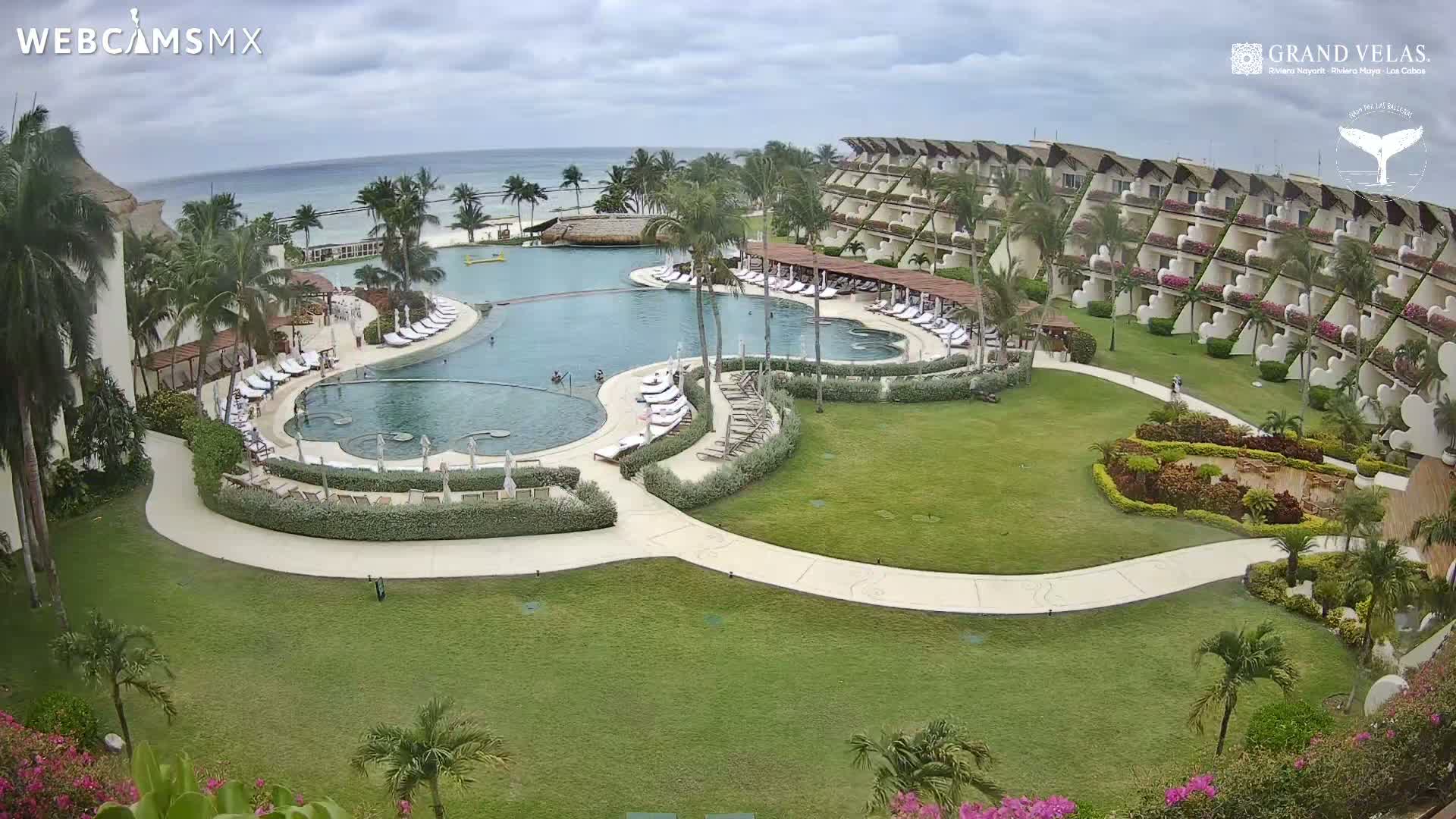 An aerial view shows a large resort pool and surrounding buildings on a partly cloudy day, overlooking a calm ocean.