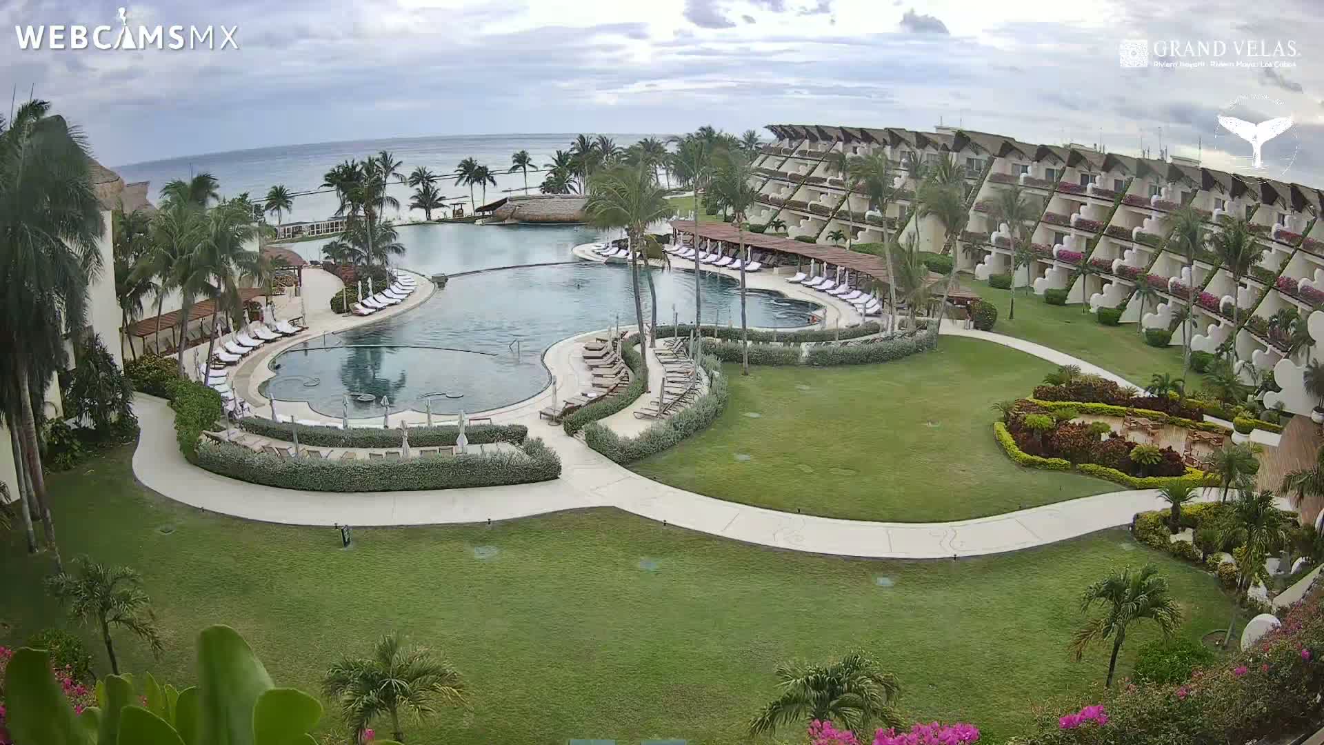 An expansive resort with a large, curved pool surrounded by lounge chairs, lush landscaping, and a multi-story building overlooks a calm ocean under a partly cloudy sky.