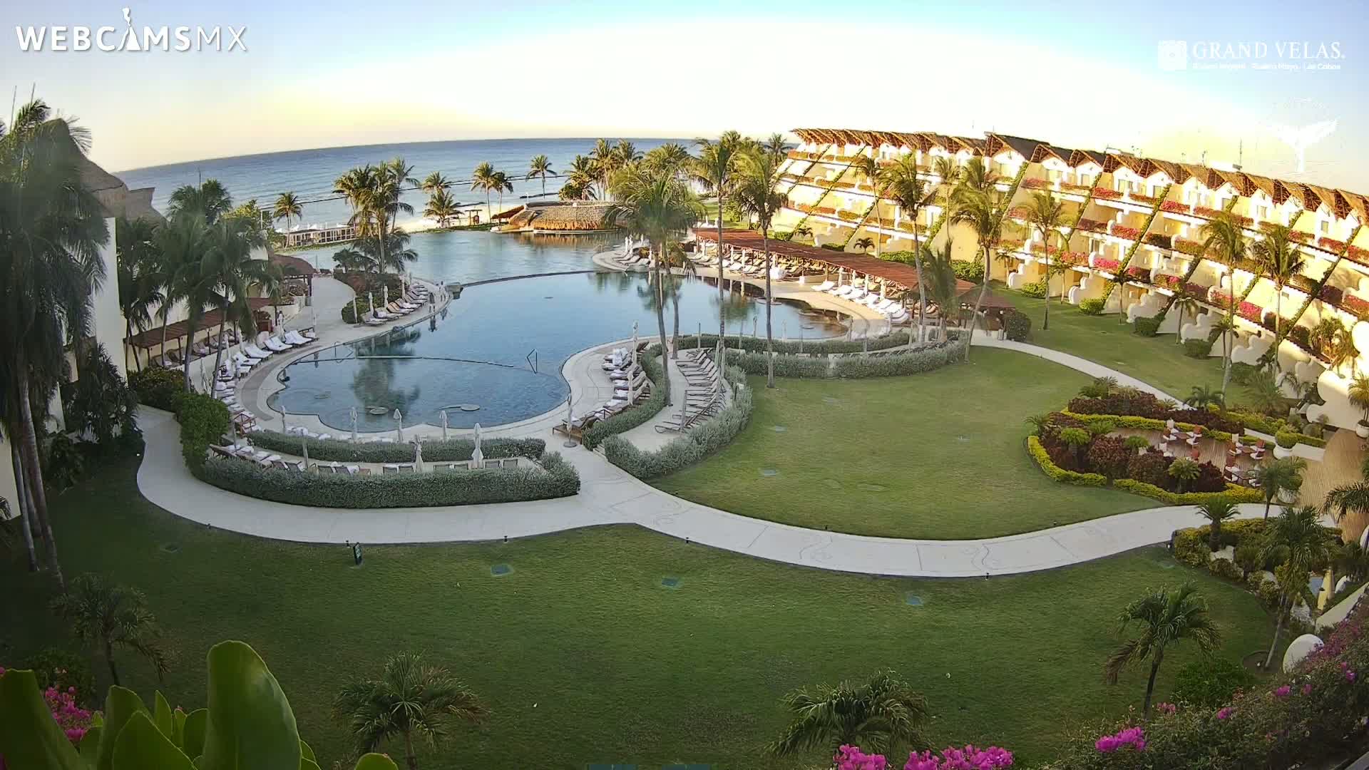 An aerial view of a resort with a large, curved pool, surrounding landscaping, and ocean in the background under a clear, sunny sky.
