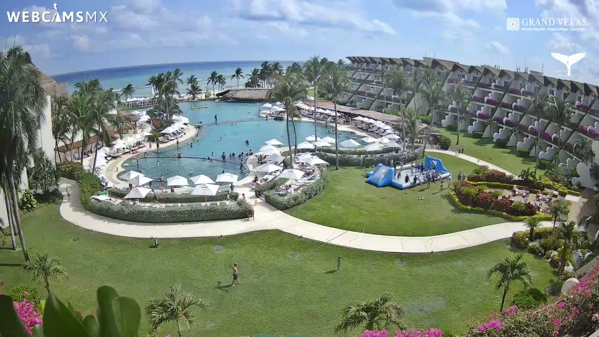 An aerial view shows a resort pool area with many people, surrounded by a large building, lush green lawns, and palm trees, under a sunny sky.