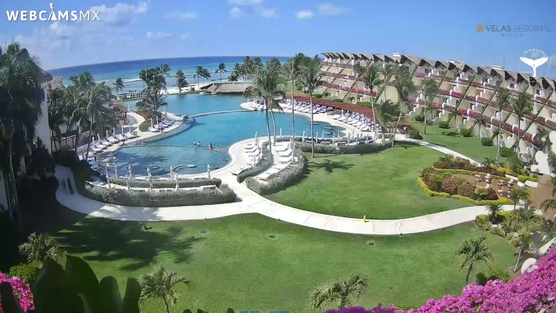 An aerial view shows a resort pool and surrounding buildings on a sunny day, with a beach and ocean visible in the background.