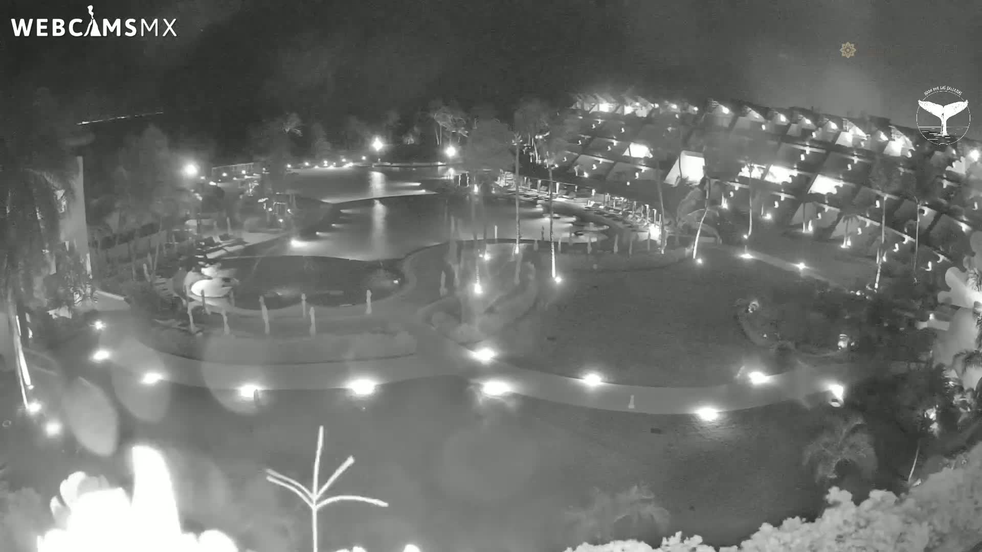 A nighttime aerial view of a resort complex with pools and surrounding buildings, illuminated by lights.