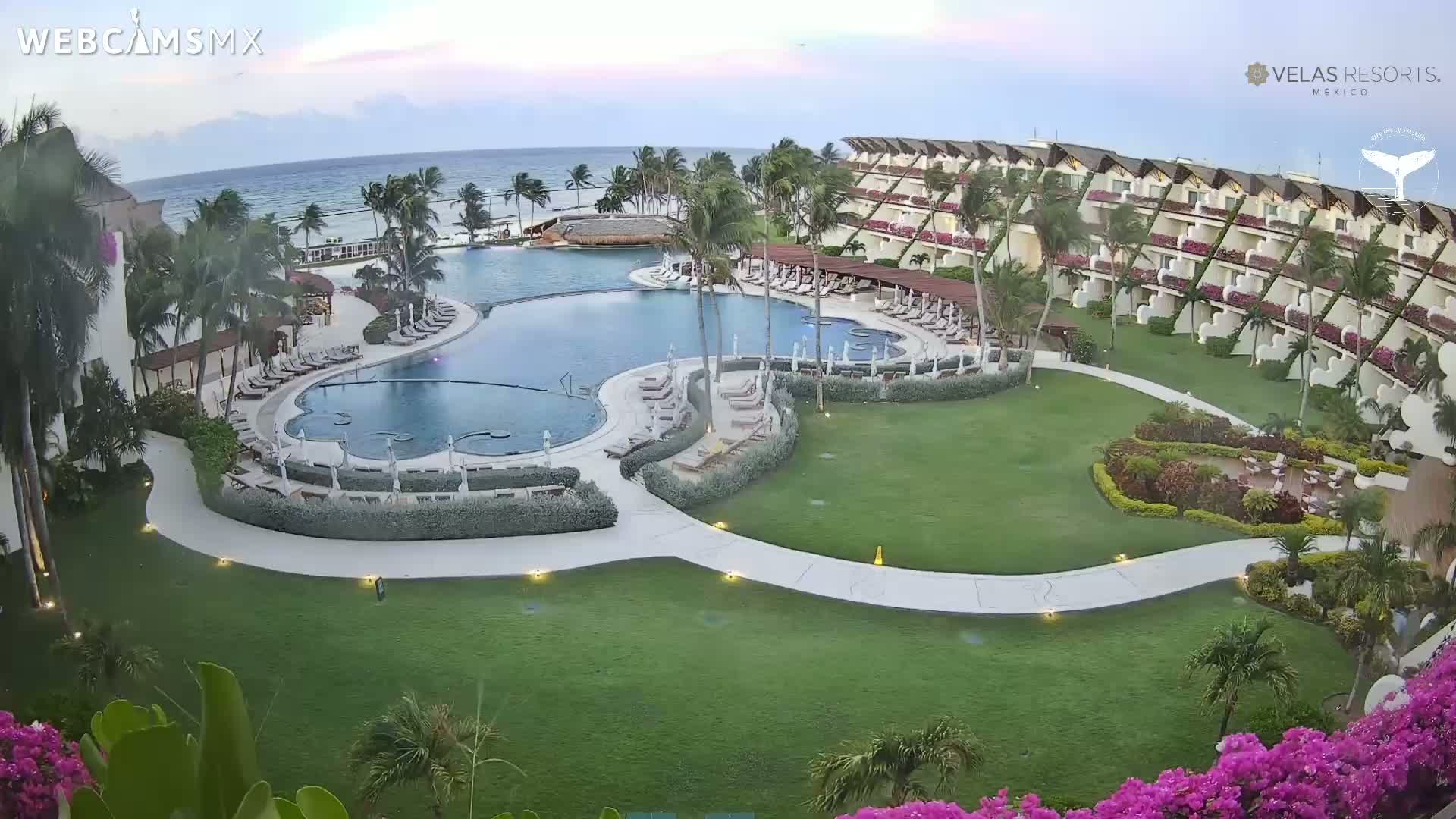 An aerial view shows a resort with a large, curved pool surrounded by lounge chairs and lush landscaping, all situated next to an ocean under a mostly clear sky.