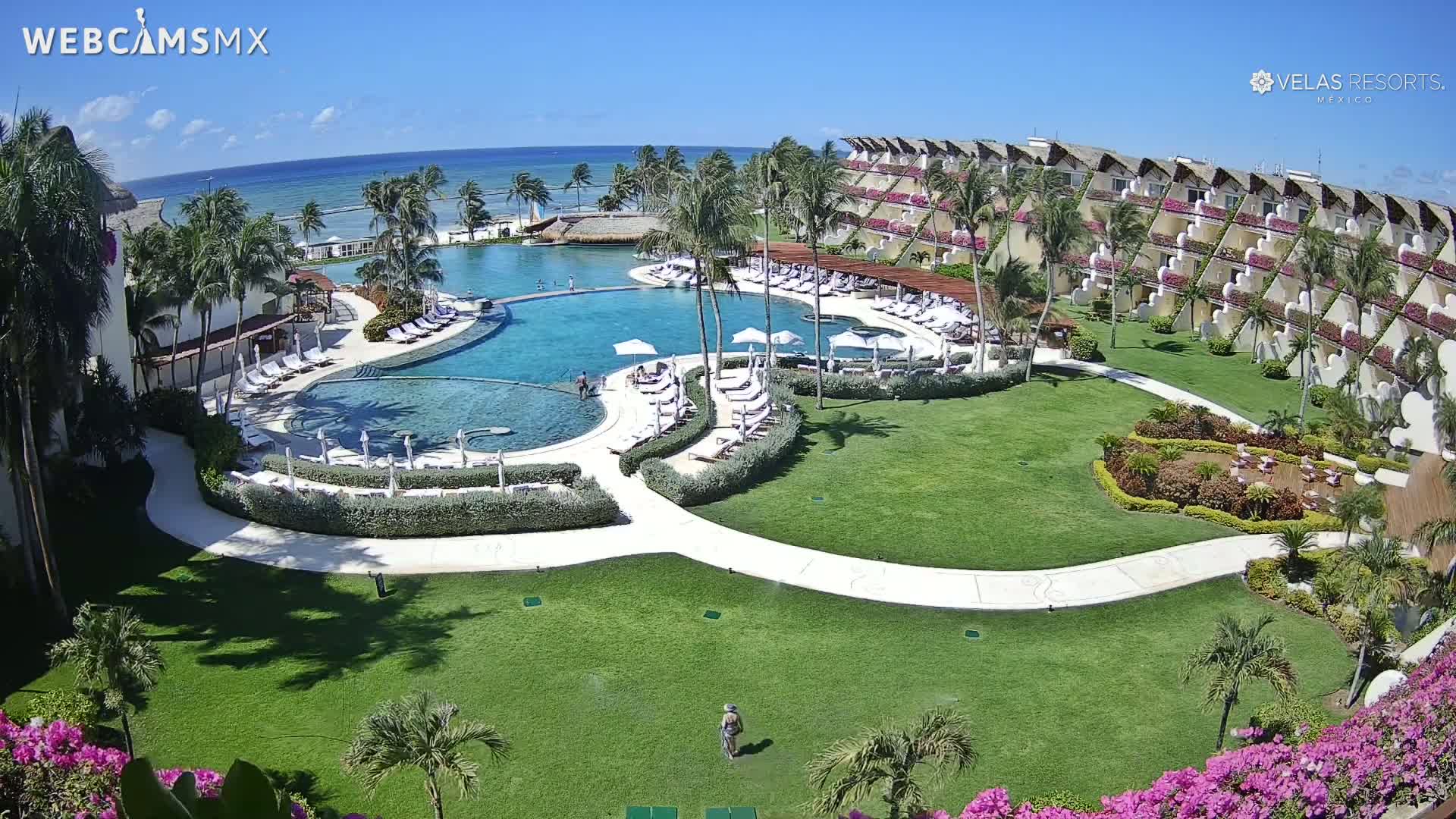 An aerial view of a resort with a large pool, surrounding landscaping, and ocean view under sunny skies.