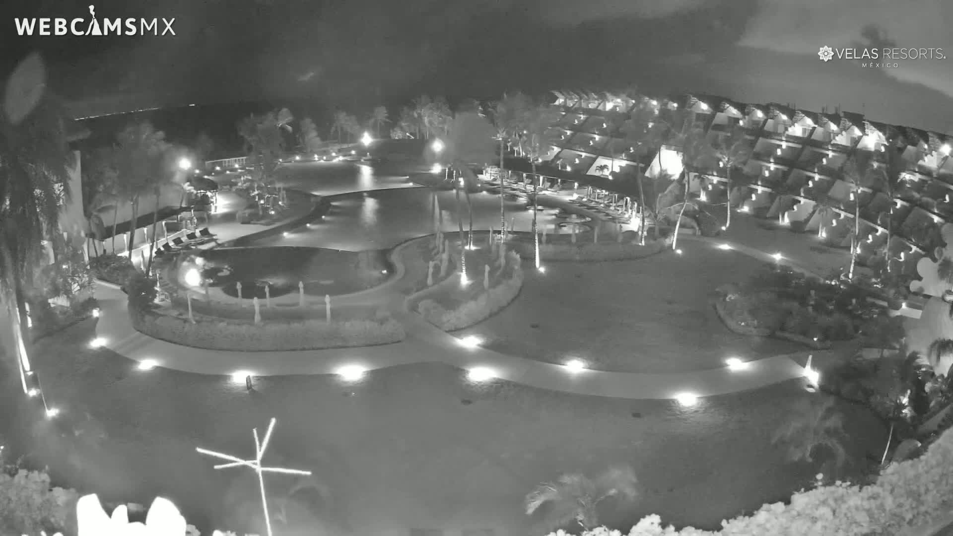 A nighttime, aerial view shows a resort with a large pool and surrounding landscaping, illuminated by lights.