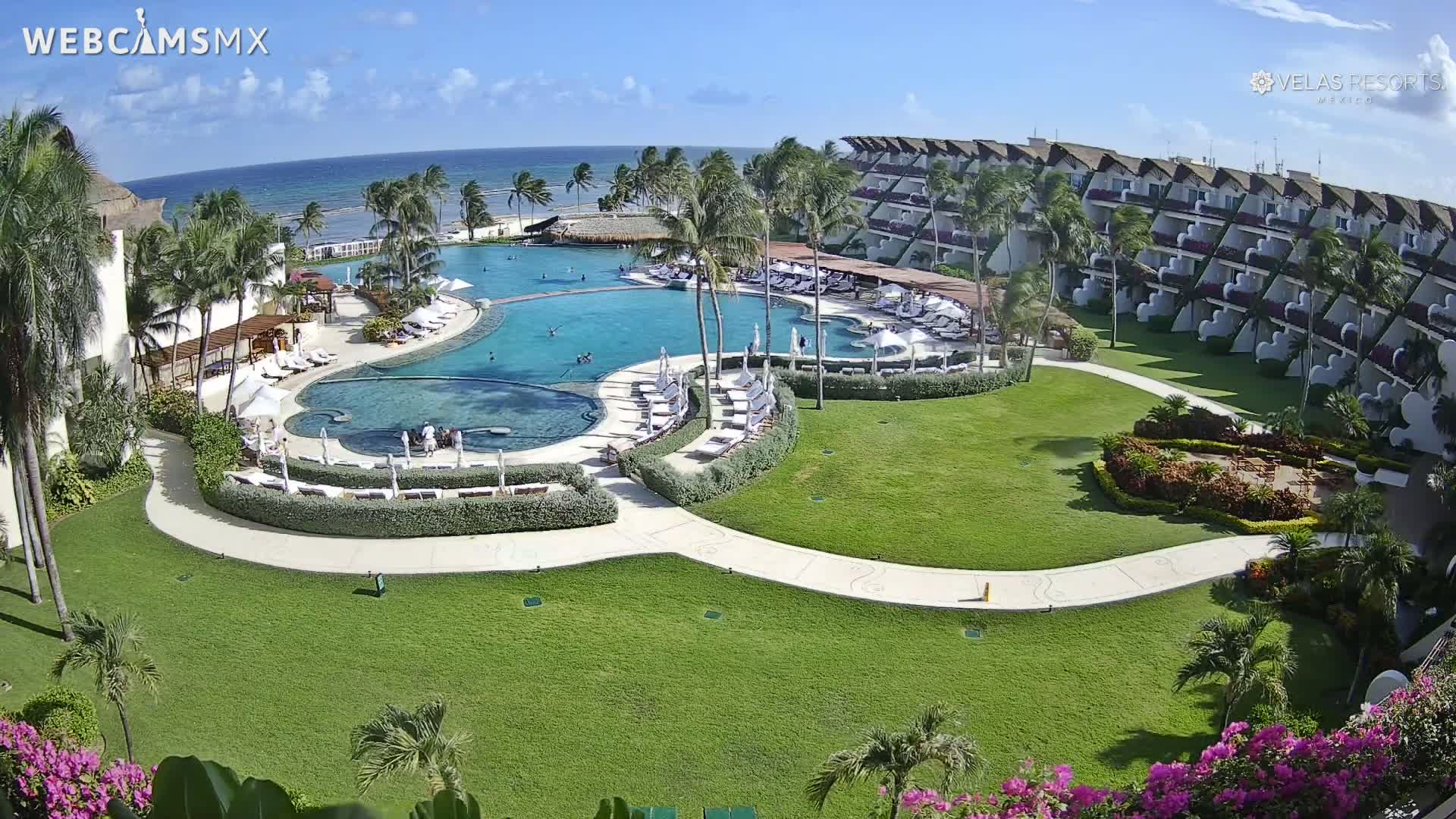 An aerial view of a resort with a large pool, surrounding landscaping, and ocean in the background on a sunny day.