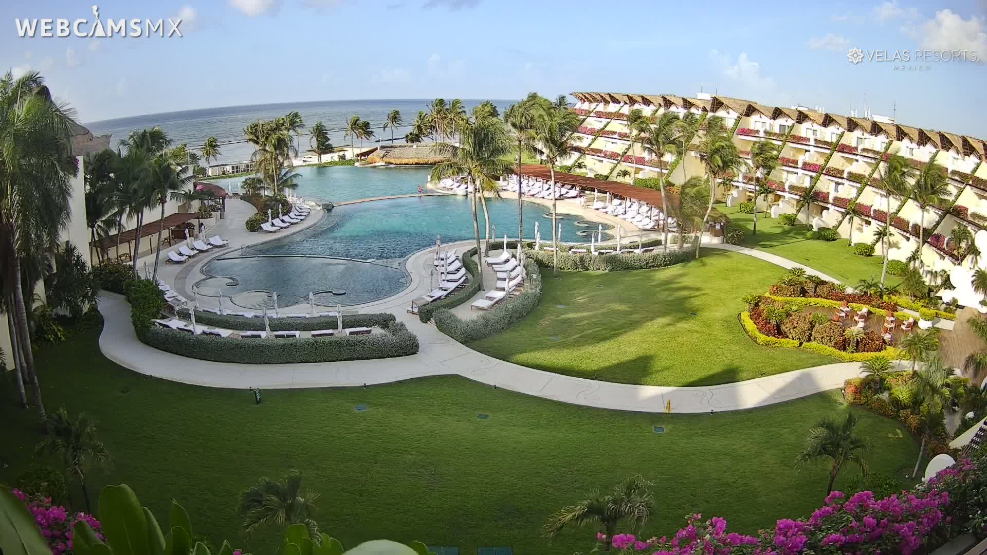 An expansive resort pool with surrounding lounge chairs and lush landscaping sits adjacent to a multi-story building, all overlooking a calm ocean under a sunny sky.