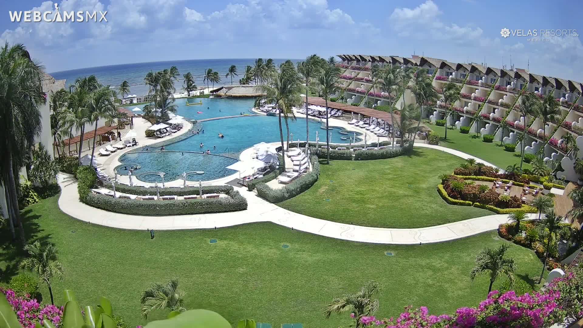 An aerial view of a resort on a sunny day shows a large, multi-level pool complex surrounded by lush green lawns, palm trees, and a multi-story building with balconies overlooking the ocean.